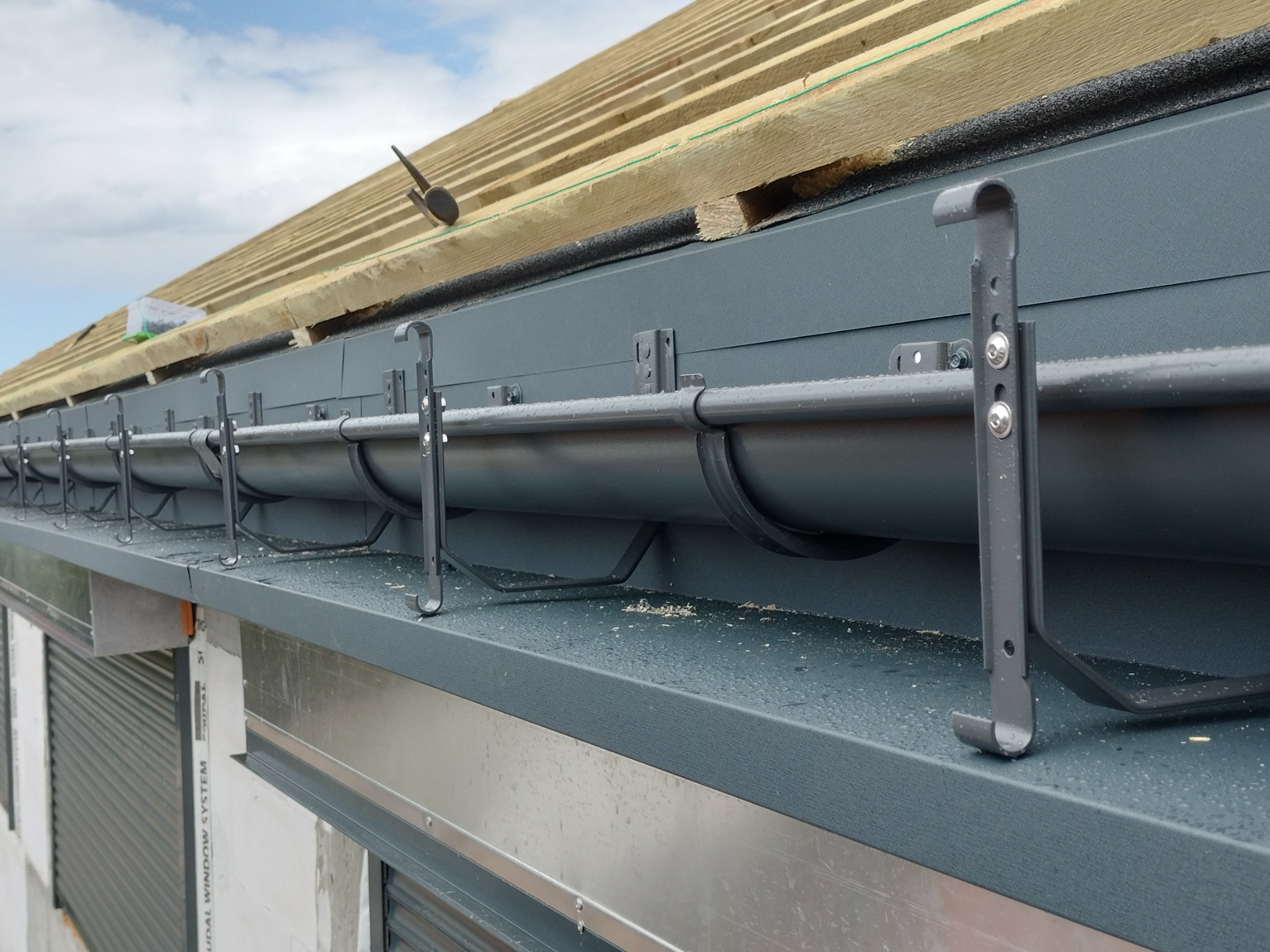 Montaż orynnowania: Szare rynny z metalowymi uchwytami zamocowane na nowym dachu z widoczną konstrukcją drewnianą, w tle błękitne niebo.