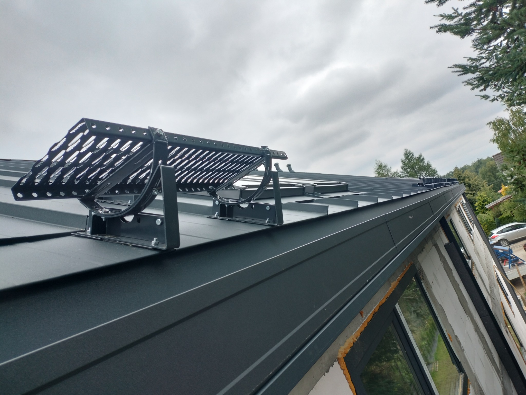 Nowoczesny, ciemnoszary dach z blachy na domu w trakcie budowy, z zamontowanym stopniem kominiarskim i widocznym ociepleniem wokół okien.