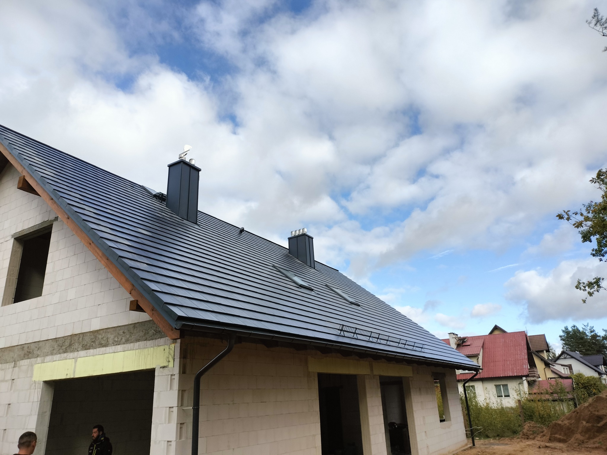 Dom w trakcie budowy, z nowym, ciemnoszarym dachem z dachówki ceramicznej, widoczne dwa kominy z metalowymi wiatrowskazami, okna dachowe, niezadaszony garaż i fragment elewacji w trakcie ocieplania.