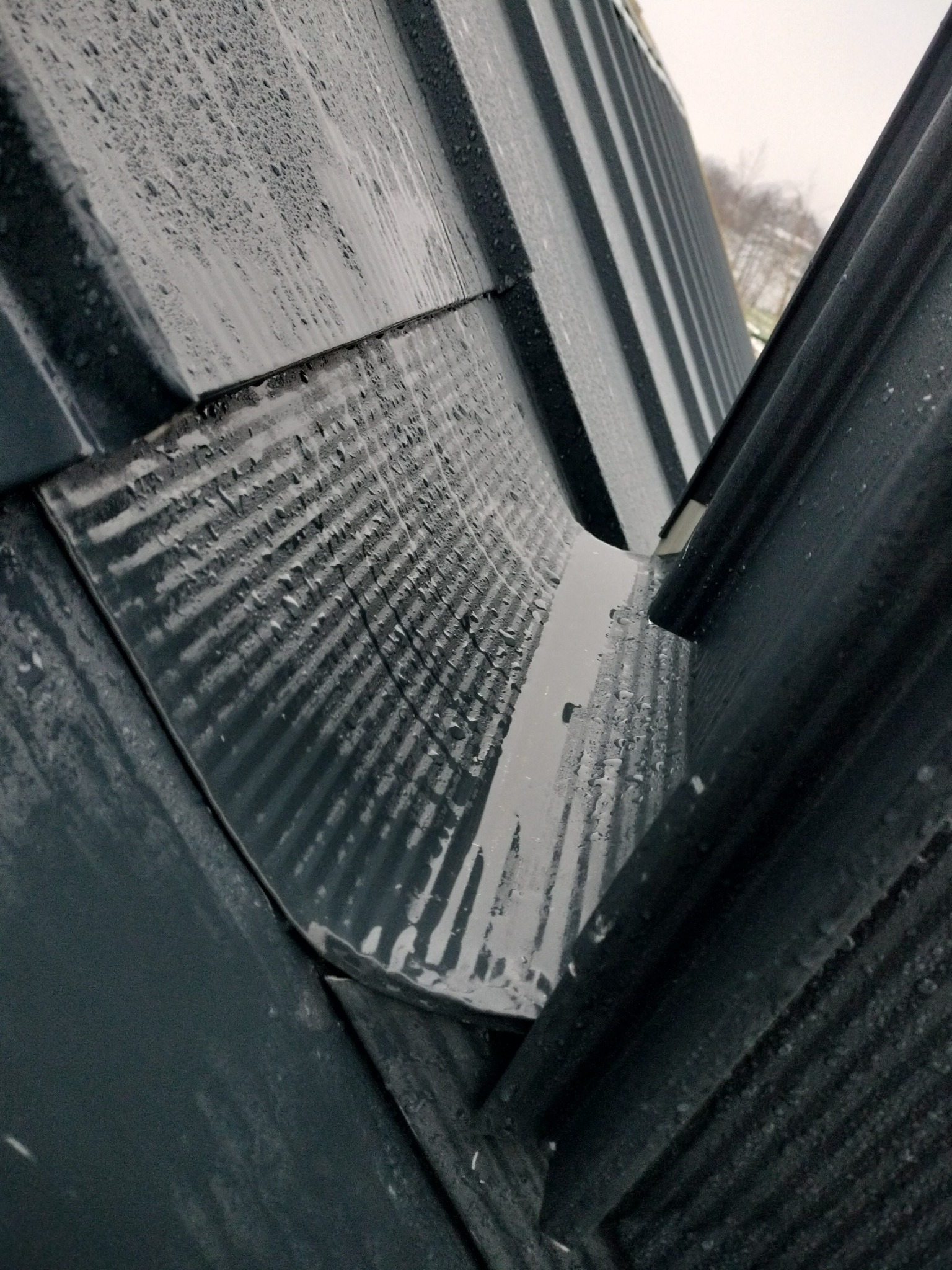 Metalowy dach pokryty kroplami deszczu, widoczny fragment rynny odprowadzającej wodę, ujęcie z dołu pod kątem.