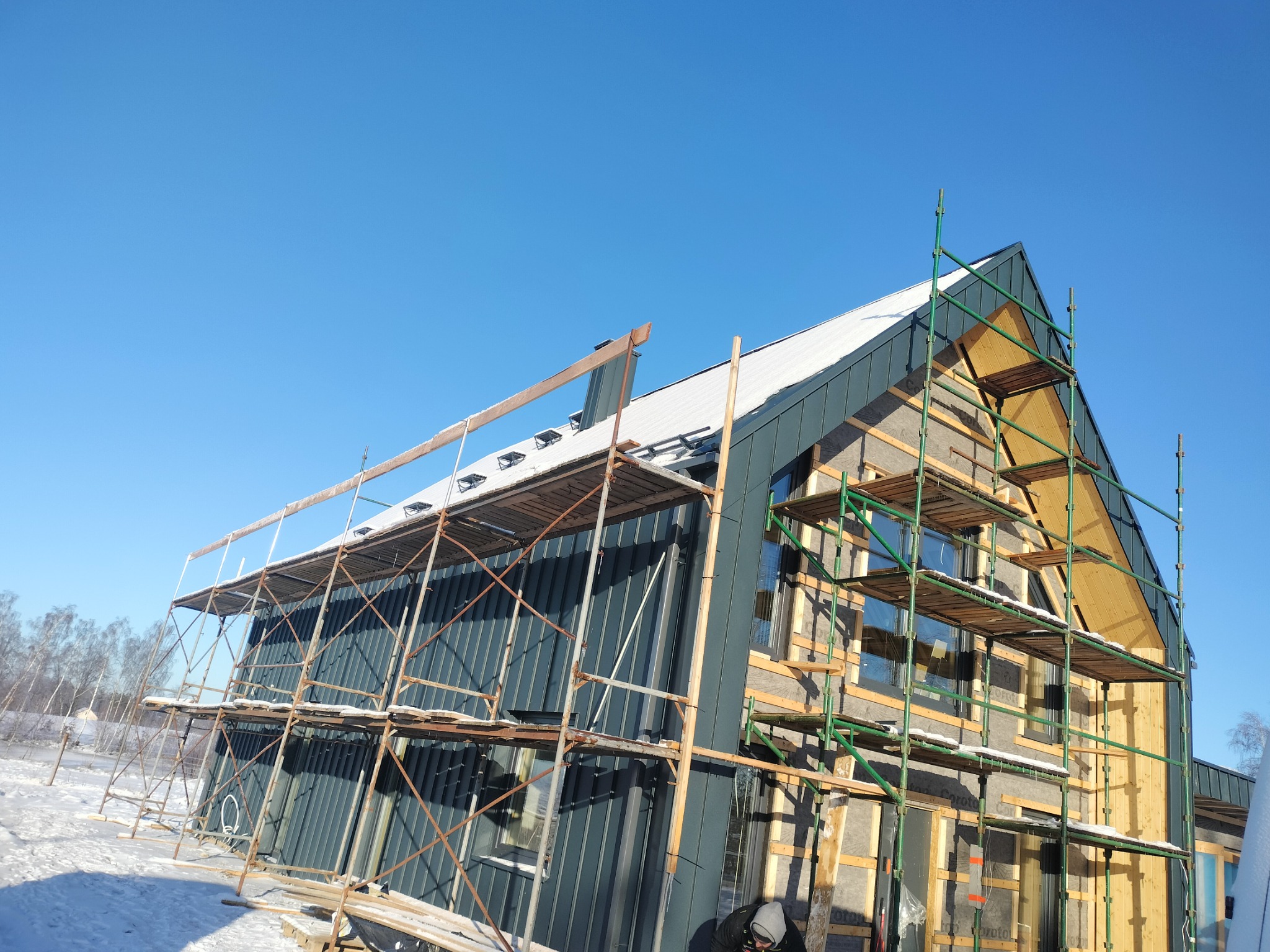 Budynek w trakcie prac wykończeniowych elewacji, widoczne rusztowania i częściowo zamontowana blacha na ścianach, ocieplenie poddasza w trakcie montażu, zimowy krajobraz w tle.