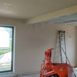 Wnętrze pokoju po nałożeniu gładzi gipsowej, widoczna maszyna tynkarska PFT G4 w kolorze pomarańczowym, aluminiowa drabina oparta o ścianę oraz wystające kable ze stropu.