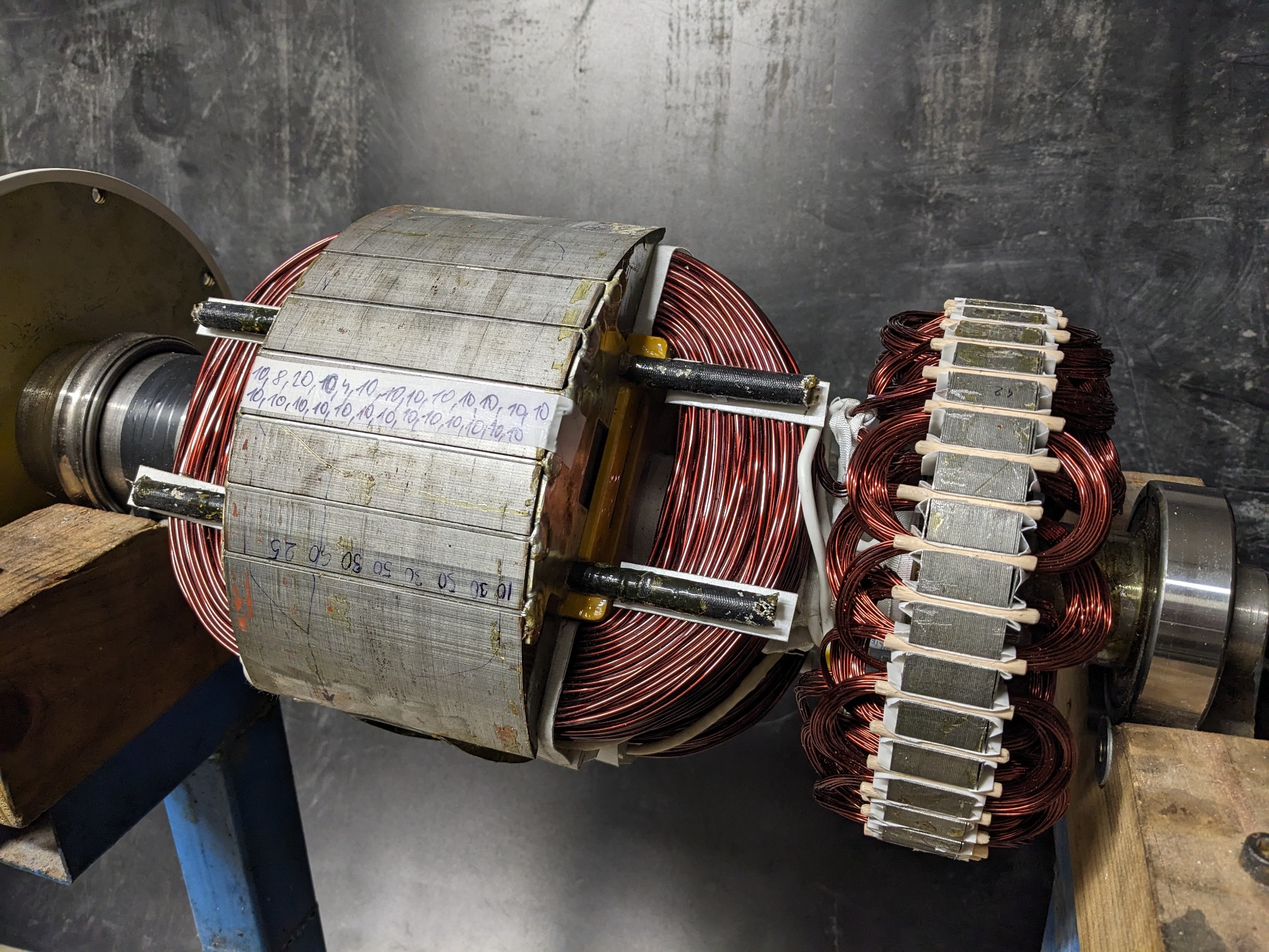 Wirnik silnika elektrycznego z uzwojeniami miedzianymi, widoczne ślady pomiarów i notatki na laminowanym rdzeniu, umieszczony na drewnianym stojaku w warsztacie.