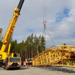 Żółty dźwig samojezdny z wysięgnikiem teleskopowym w trakcie montażu, na żwirowym placu budowy, w tle las iglasty i pochmurne niebo.