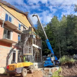 Budowa domu z cegły, widoczna konstrukcja szalunkowa, pracownicy na rusztowaniu, żółta ładowarka teleskopowa Mecalac i niebieski dźwig na tle drzew i błękitnego nieba.