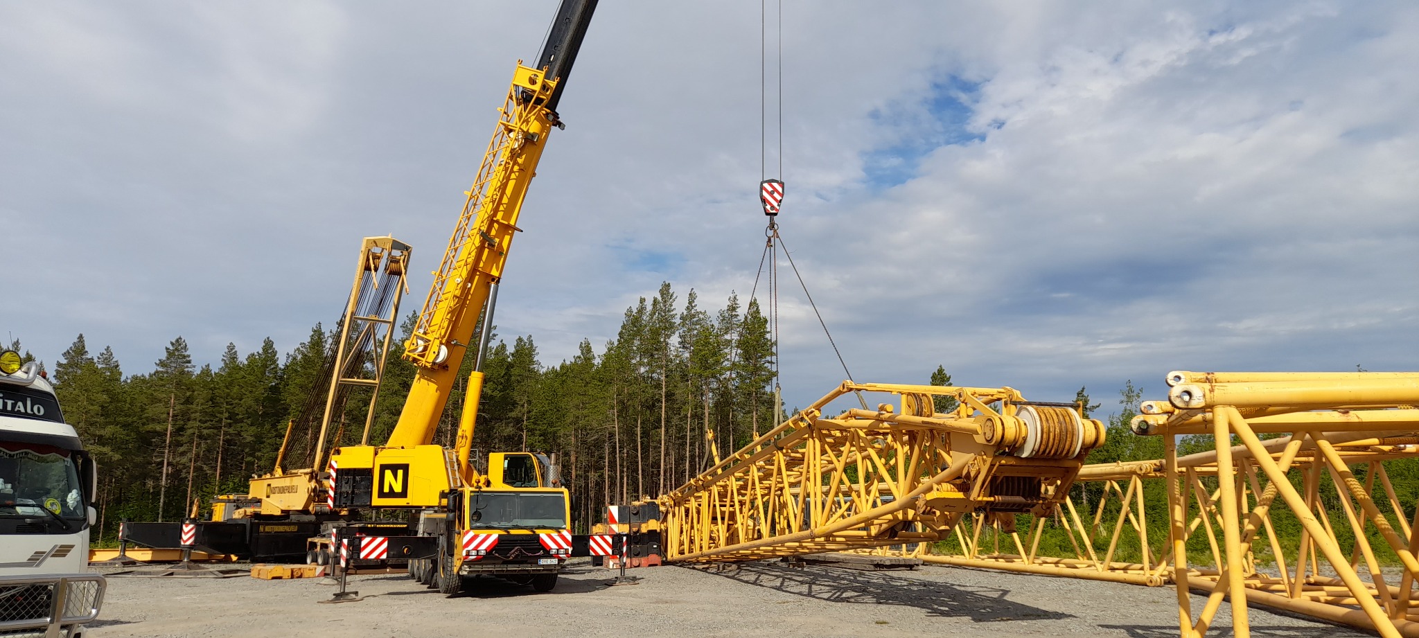 Żółty dźwig samojezdny z wysięgnikiem teleskopowym w trakcie montażu, na żwirowym placu budowy, w tle las iglasty i pochmurne niebo.