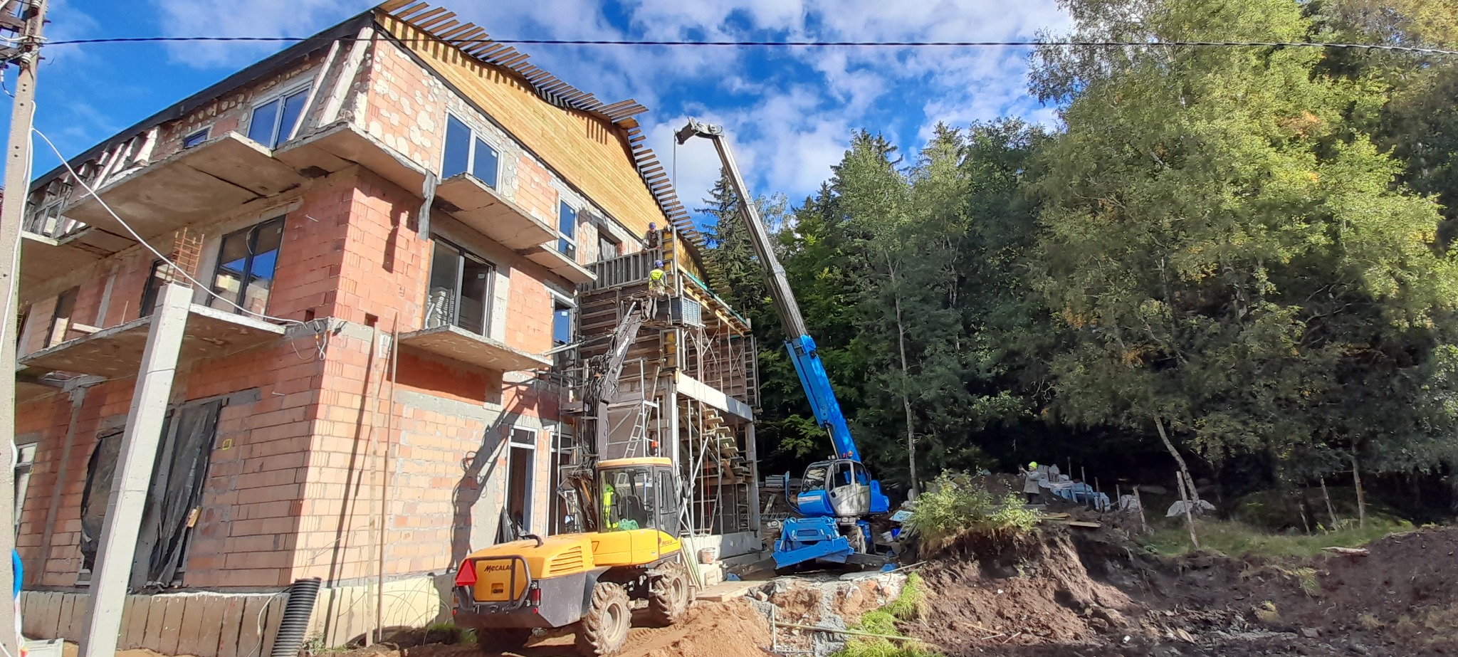 Budowa domu z cegły, widoczna konstrukcja szalunkowa, pracownicy na rusztowaniu, żółta ładowarka teleskopowa Mecalac i niebieski dźwig na tle drzew i błękitnego nieba.