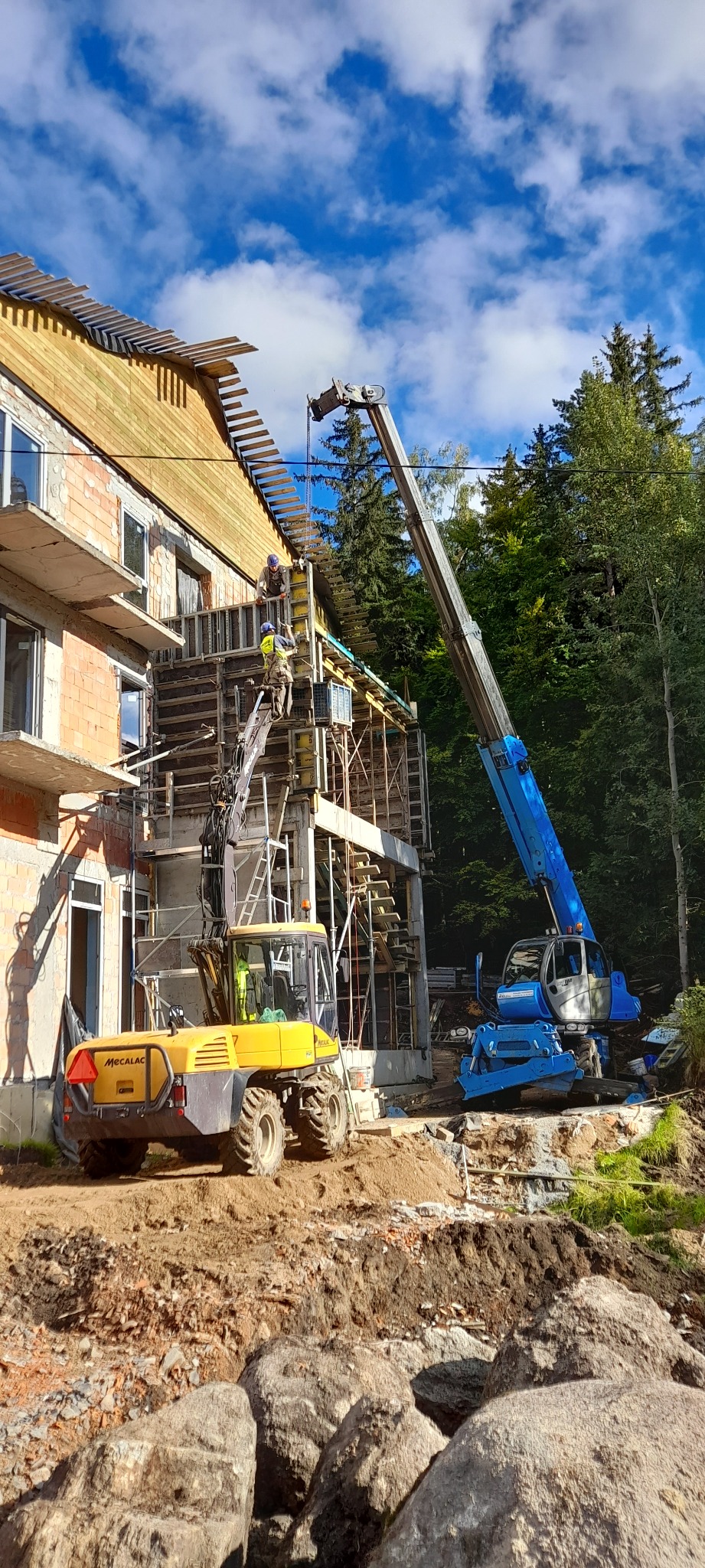 Żółta koparka Mecalac i niebieski dźwig na placu budowy, pracownicy montują szalunki na ścianach budynku w stanie surowym otwartym, w tle las i błękitne niebo z chmurami.