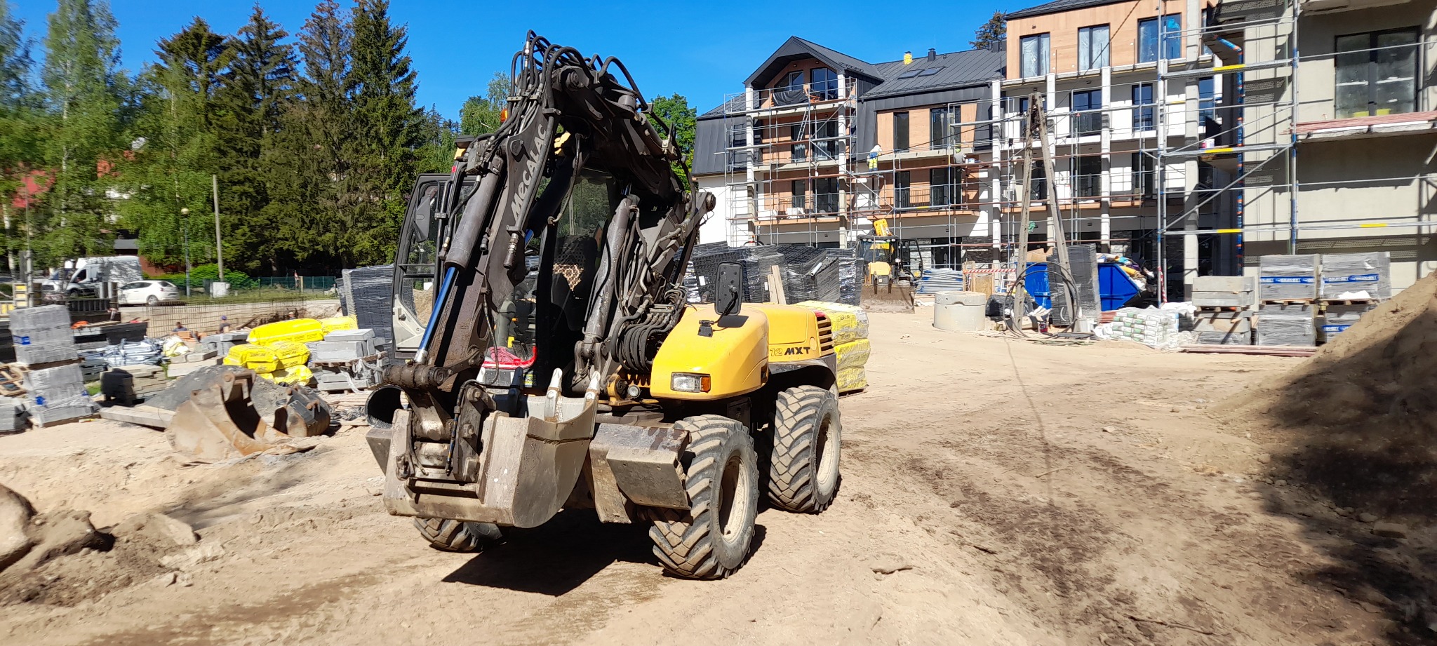 Żółta koparka Mecalac 12 MXT na placu budowy z nowym budynkiem w tle, stosy materiałów budowlanych, ziemia i drzewa.