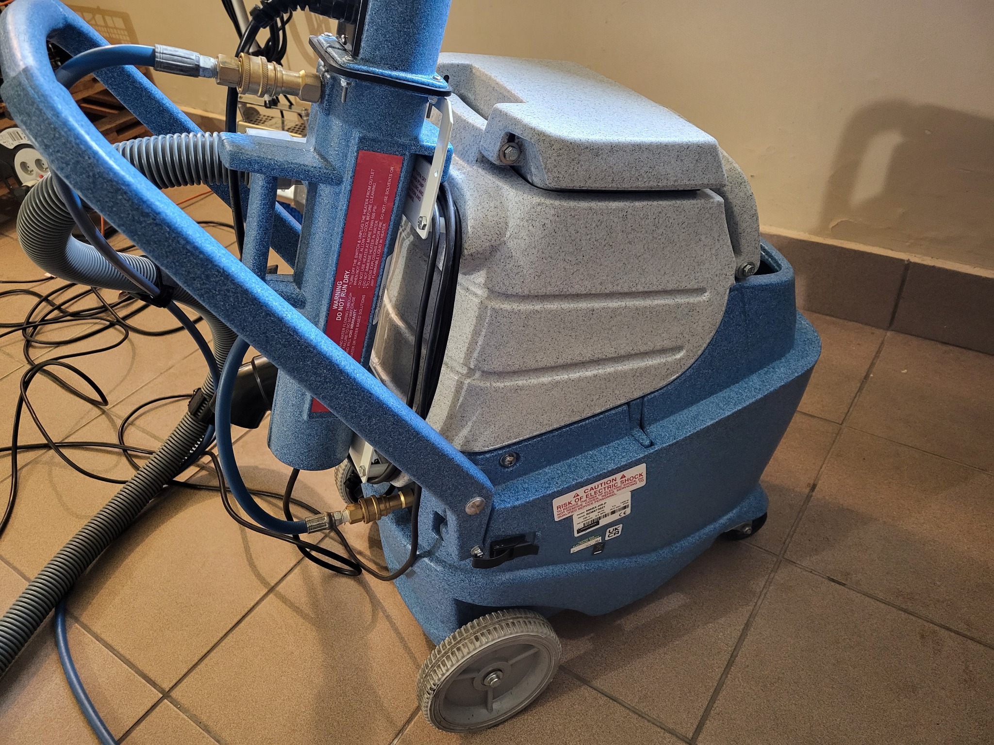 Profesjonalna, niebiesko-szara maszyna do czyszczenia dywanów i tapicerki, z widocznymi wężami i kablami, stojąca na kafelkach podłogowych.