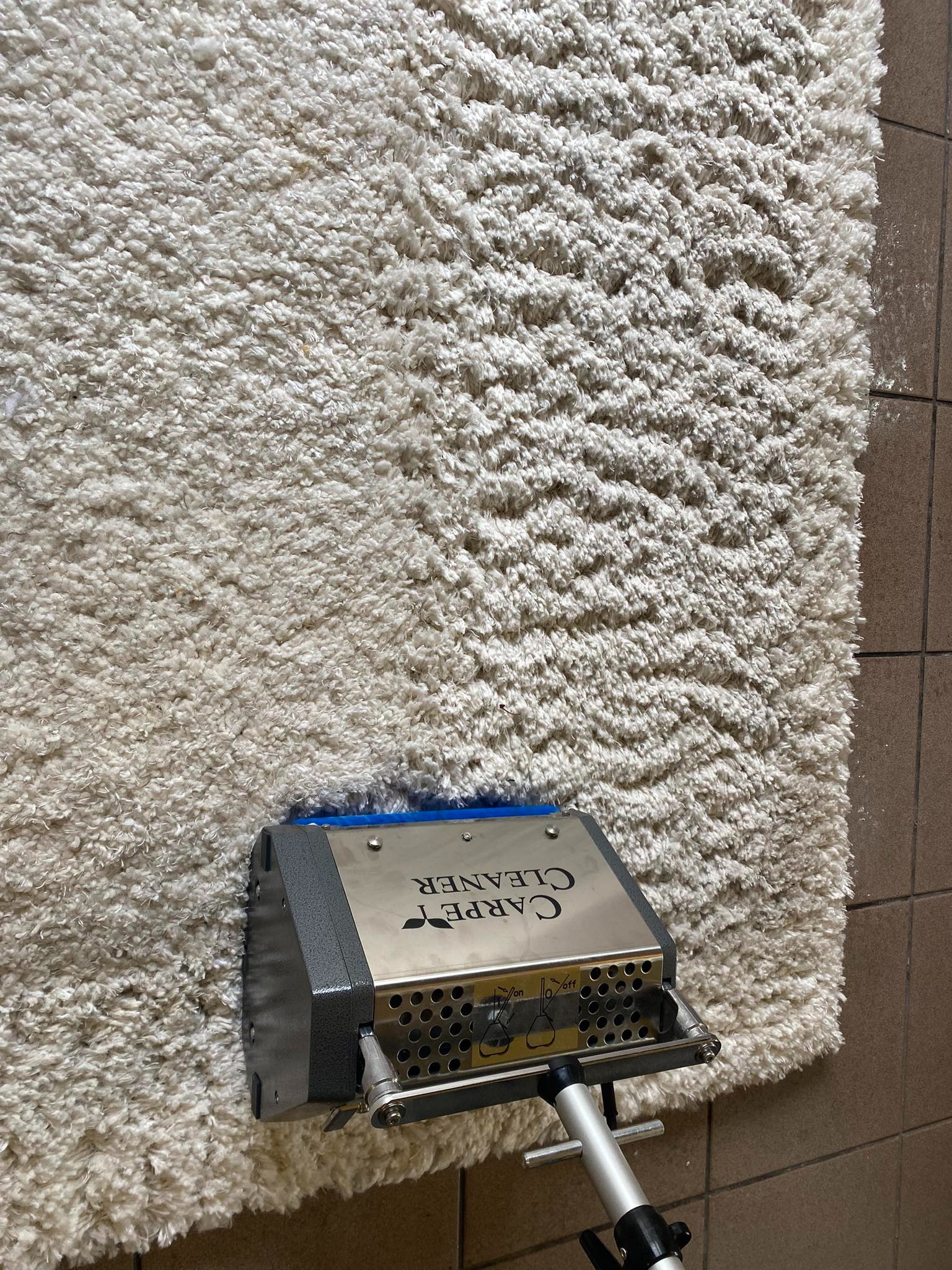Profesjonalny odkurzacz piorący z napisem 'CARPET CLEANER' w trakcie czyszczenia jasnego, puszystego dywanu na tle brązowych płytek podłogowych.