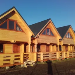 Rząd identycznych, świeżo wybudowanych drewnianych domków z ciemnymi, spadzistymi dachami i małymi balkonami, oświetlonych popołudniowym słońcem.