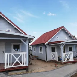 Dwa małe, szare domki z czerwonymi dachami i białymi balustradami, połączone ścieżką z kostki brukowej, na etapie wykończenia terenu wokół nich.