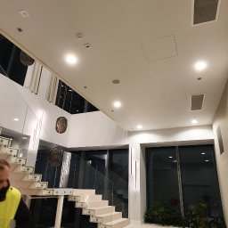 Ihor Panasiuk - Wnętrze przestronnego holu z białymi schodami ze szklaną balustradą, widoczne fragmenty sufitu z kratką wentylacyjną i otworem rewizyjnym oraz pracownik w odblaskowej kamizelce z wiadrem przy...