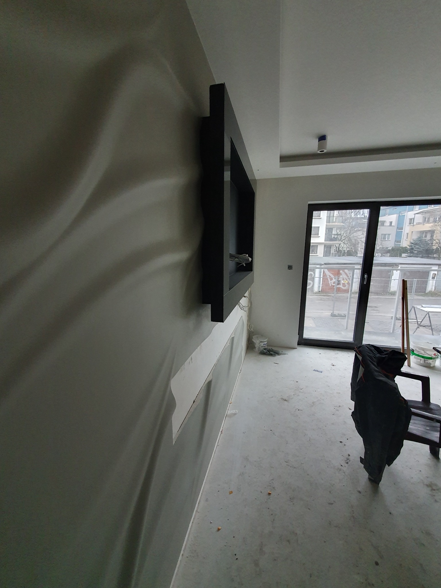 Ściana z dekoracyjną fakturą, częściowo zakryta taśmą malarską, obok czarna półka wnękowa, widok na okno balkonowe i elementy wyposażenia wnętrza w trakcie prac wykończeniowych.