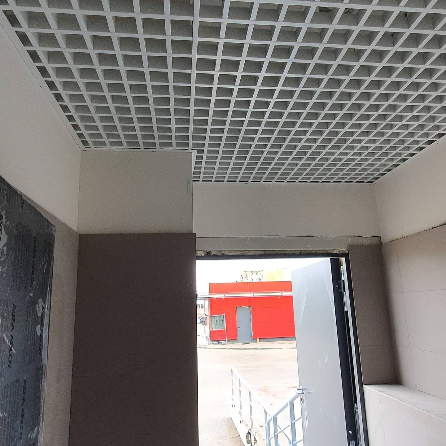 Kwadratowa, biała kratownica sufitowa w biurze, widok z dołu, z otwartymi drzwiami i widokiem na czerwony budynek na zewnątrz.