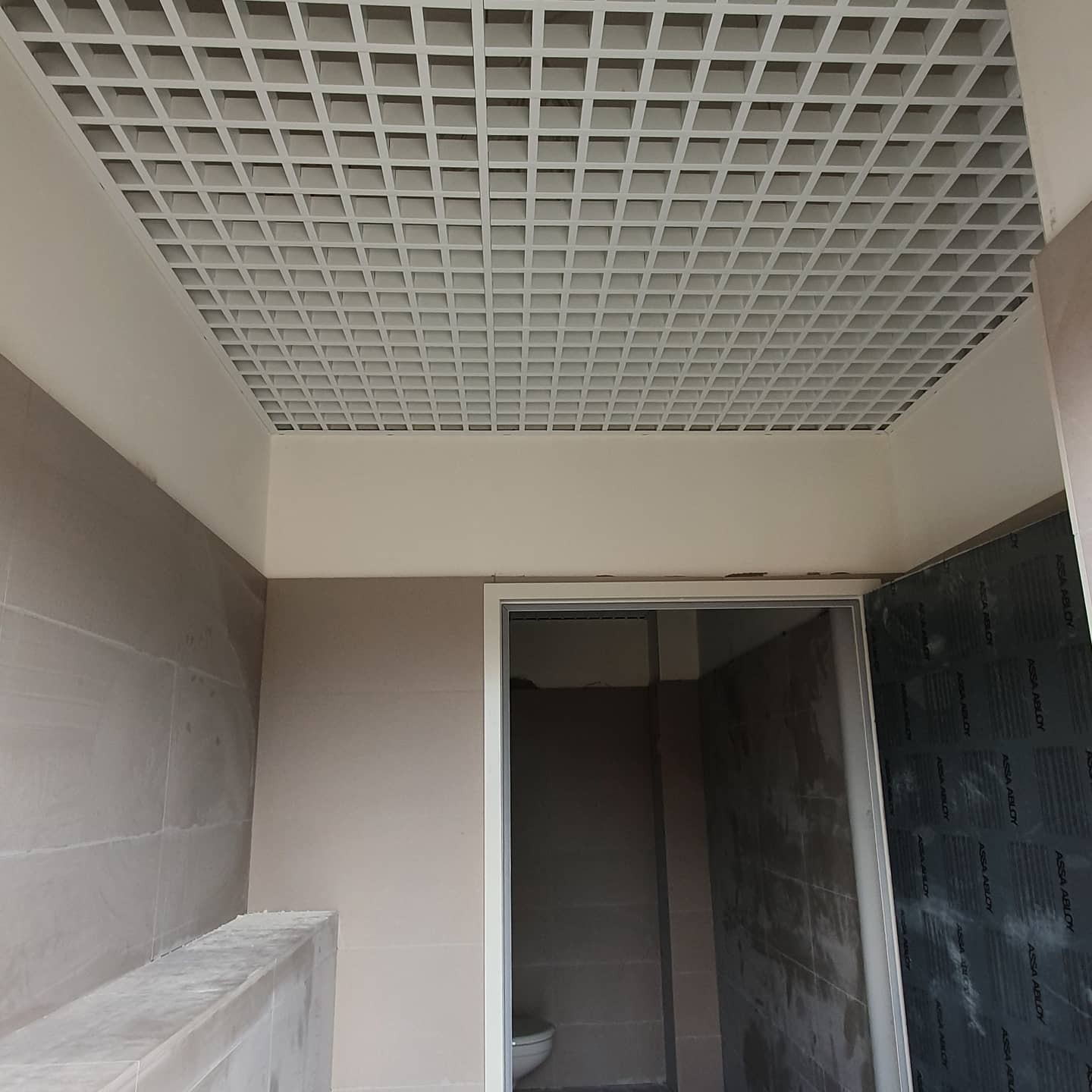 Kasetonowy sufit podwieszany z kratką w łazience w trakcie remontu, widok na częściowo wykończone pomieszczenie z widocznym fragmentem toalety i zabezpieczoną folią ścianą.
