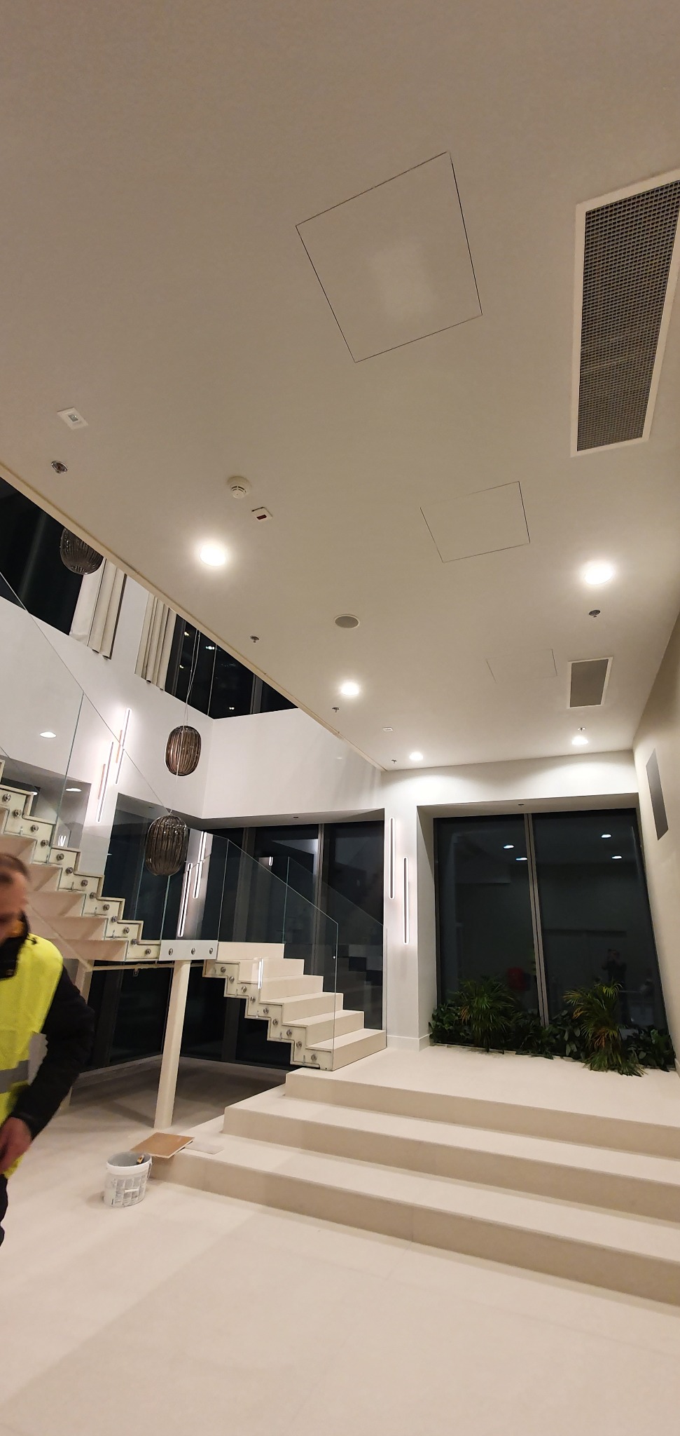 Wnętrze przestronnego holu z białymi schodami ze szklaną balustradą, widoczne fragmenty sufitu z kratką wentylacyjną i otworem rewizyjnym oraz pracownik w odblaskowej kamizelce z wiadrem przy...