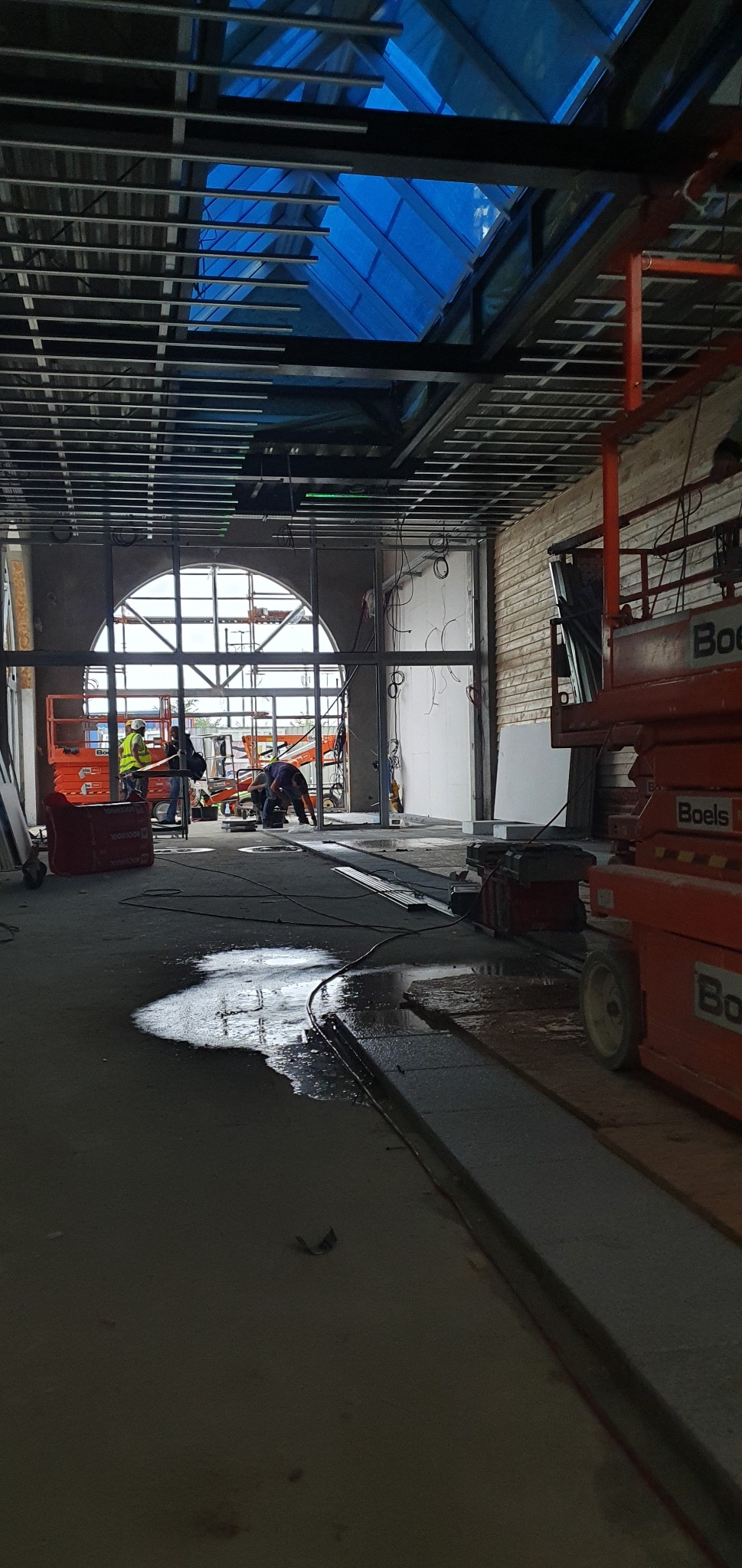 Wnętrze w trakcie remontu: widoczne rusztowania sufitowe, otwór okienny w kształcie półkola, pracownicy na podnośnikach i kałuża wody na podłodze.