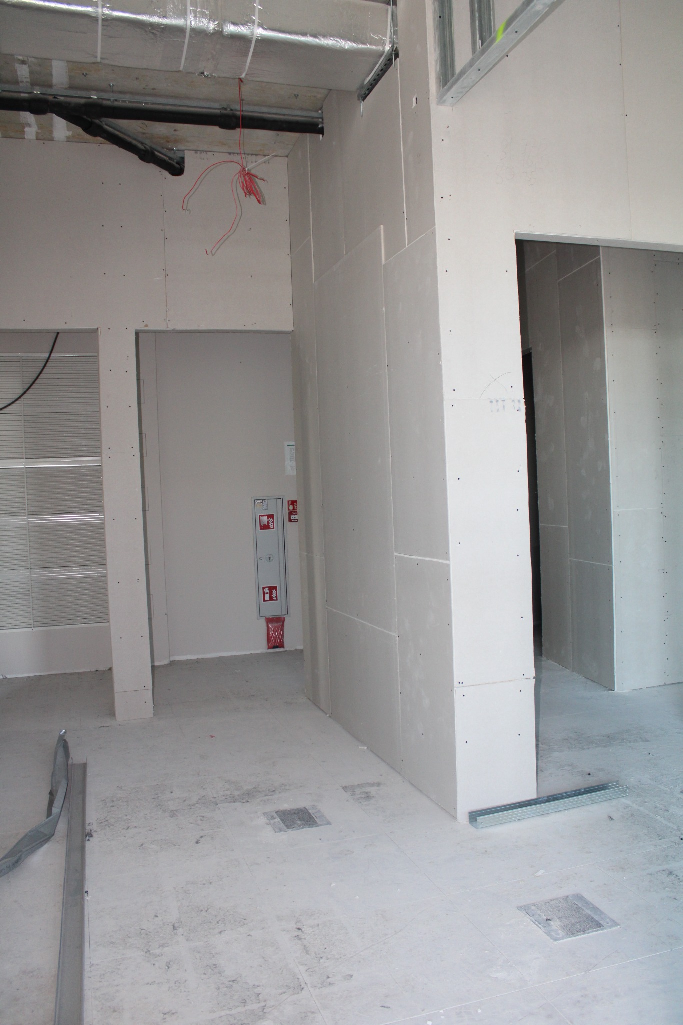 Wnętrze w trakcie prac wykończeniowych: surowe ściany z płyt gipsowo-kartonowych, widoczne profile metalowe, instalacje pod sufitem, posadzka z szarych płytek. W tle skrzynka z zabezpieczeniami...