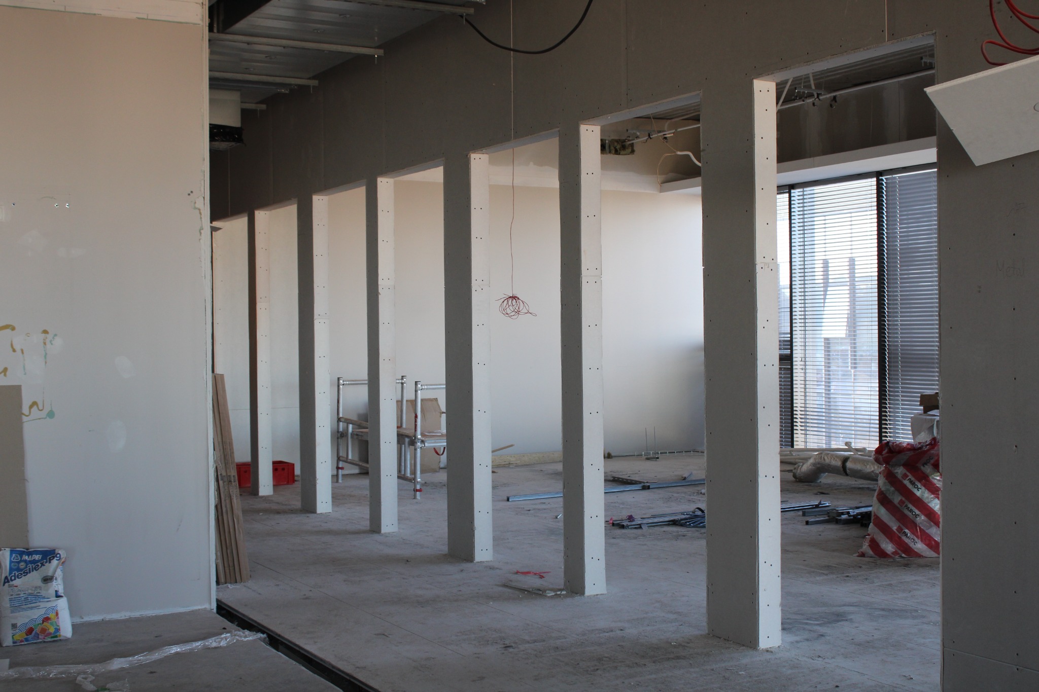 Wnętrze w trakcie prac wykończeniowych z widocznymi słupkami z płyt gipsowo-kartonowych, przewodami zwisającymi z sufitu i elementami instalacji. W tle widoczne okna z żaluzjami.