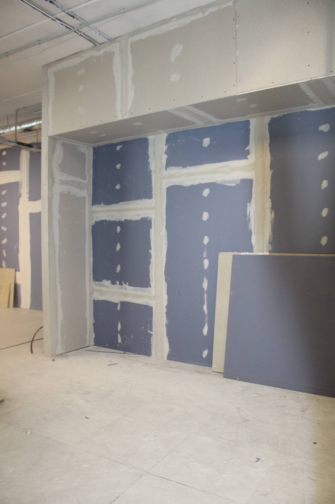 Niedokończona konstrukcja ścian działowych z płyt gipsowo-kartonowych w szarym pomieszczeniu, widoczne spoiny wypełnione masą szpachlową, płyty czekają na wykończenie.