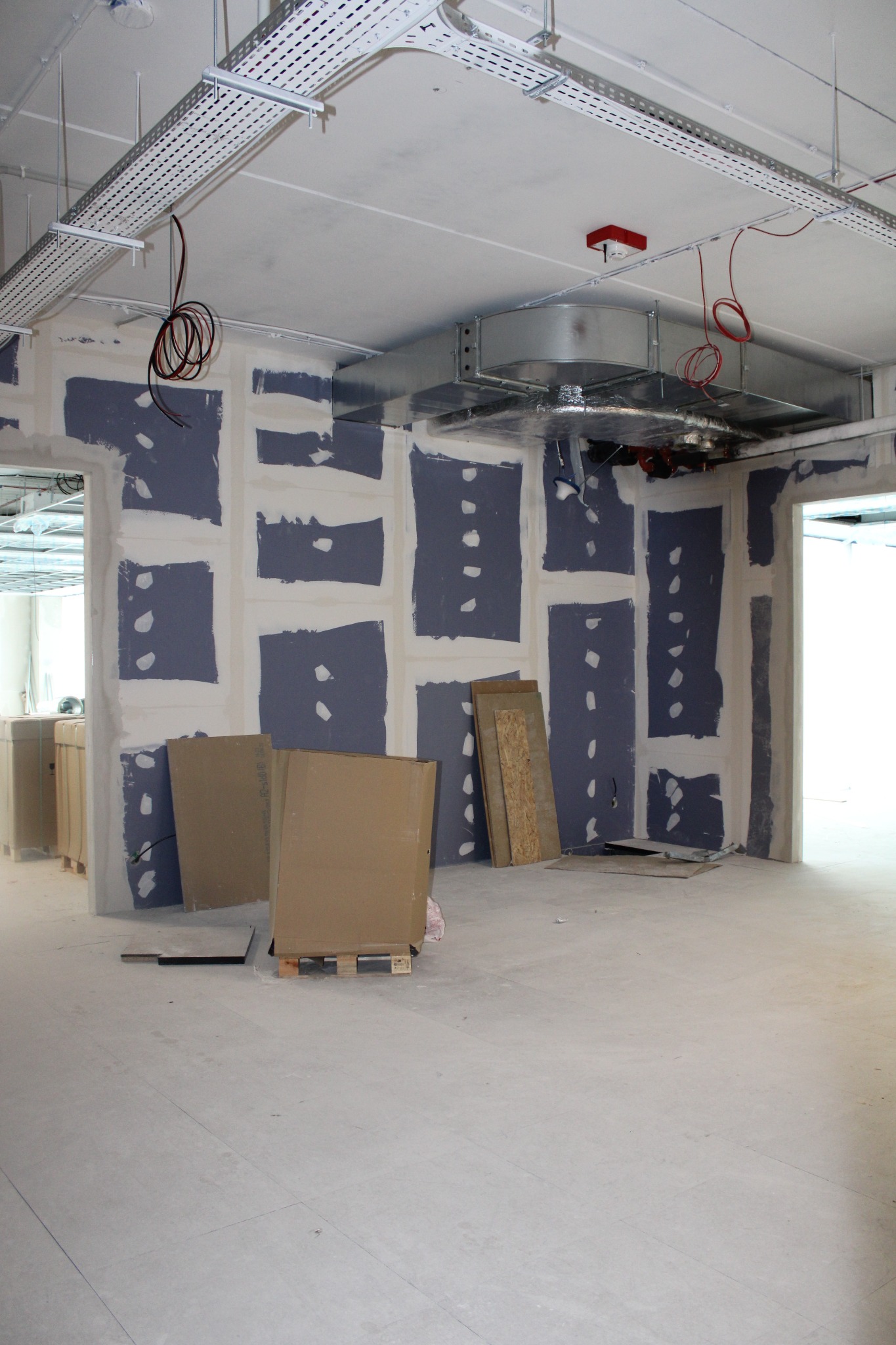 Wnętrze w trakcie prac wykończeniowych: ściany z widocznymi fragmentami płyt, częściowo pomalowane, instalacja wentylacyjna pod sufitem, kable zwisające z sufitu, płyty budowlane oparte o ścianę.