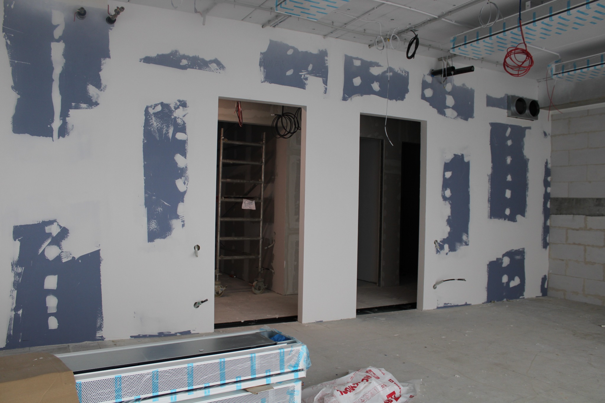 Ściana z nałożonymi łatami gipsowymi przed szlifowaniem, widoczne dwa otwory drzwiowe, instalacje pod sufitem i materiały budowlane na podłodze.