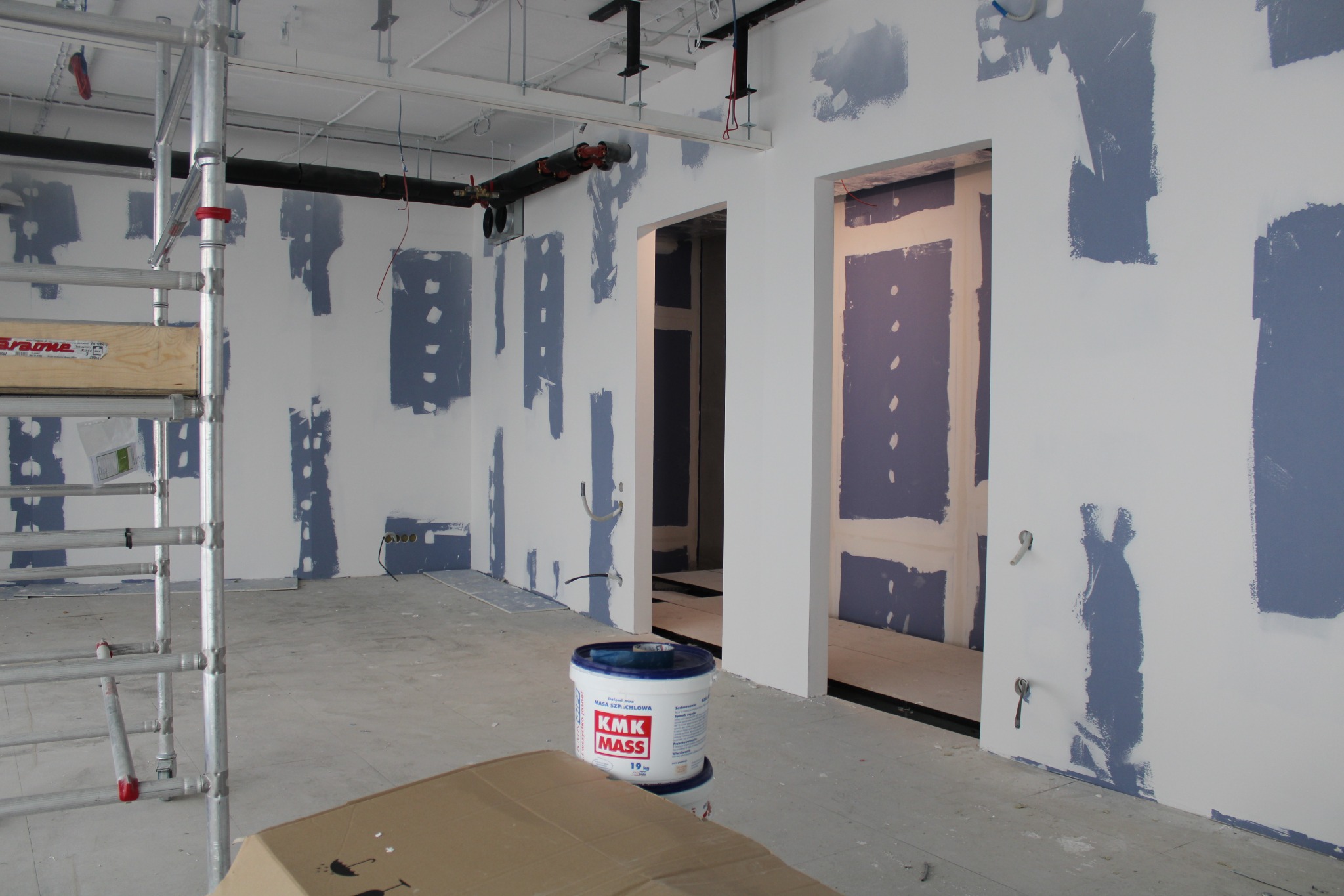Wnętrze w trakcie przygotowania do malowania: ściany z widocznymi miejscami szpachlowania, wiadro z masą szpachlową na pierwszym planie, widoczne instalacje pod sufitem i fragment rusztowania.