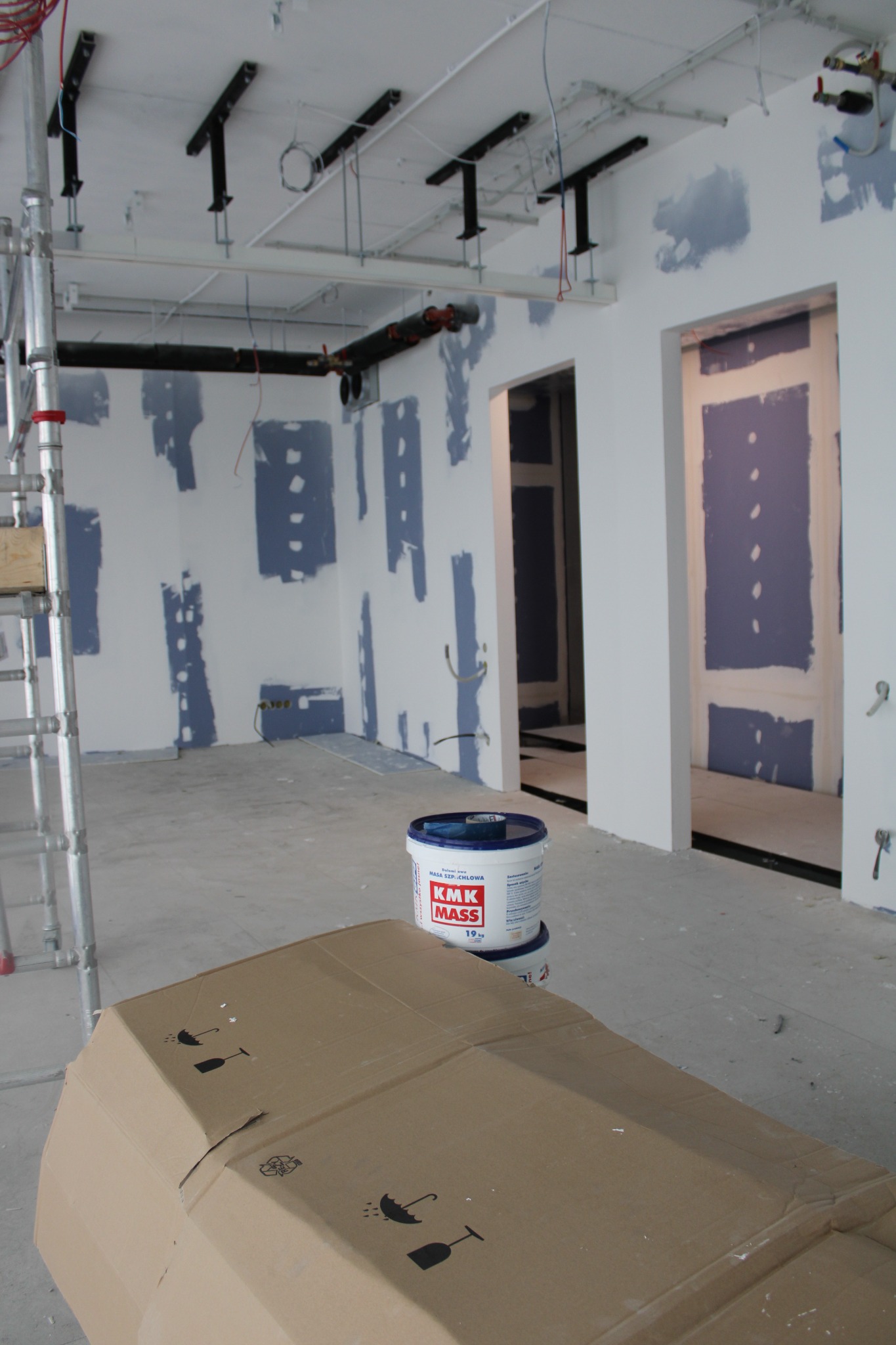 Wnętrze w trakcie prac wykończeniowych: ściany z fragmentami nałożonej gładzi, widoczne otwory drzwiowe, puszka z masą szpachlową i kartonowe pudło na pierwszym planie, rusztowanie z lewej strony.