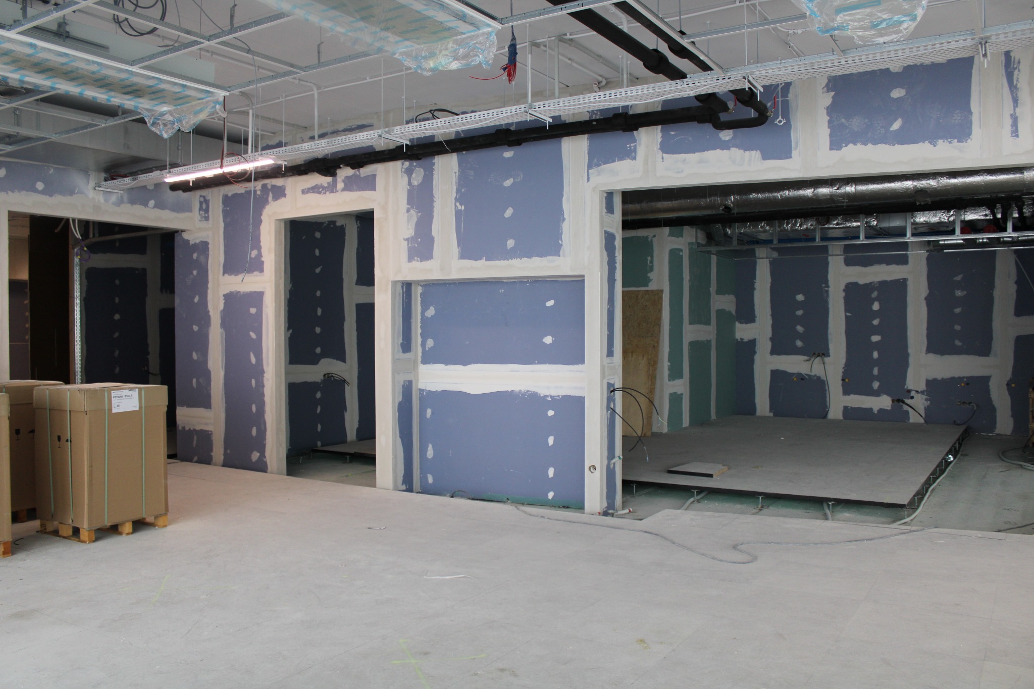 Wnętrze biurowe w trakcie prac wykończeniowych: ściany z płyt gipsowo-kartonowych w kolorze niebieskim, częściowo zaszpachlowane, widoczne instalacje pod sufitem, kartony z materiałami budowlanymi...