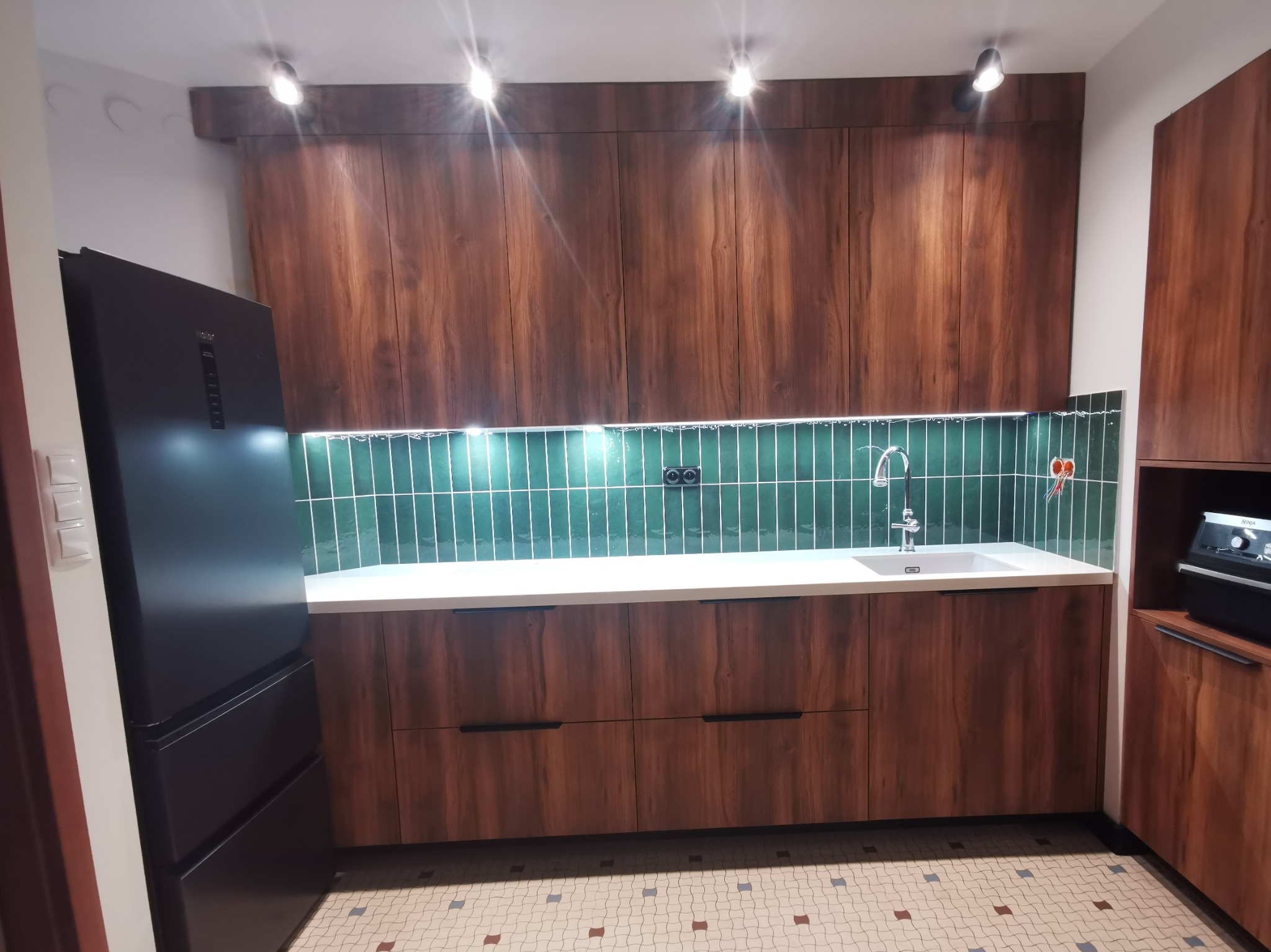 Nowoczesna kuchnia z drewnopodobnymi frontami w odcieniu dębu barokowego bursztynowego, białym blatem, zielonymi płytkami na ścianie i czarną lodówką side-by-side.