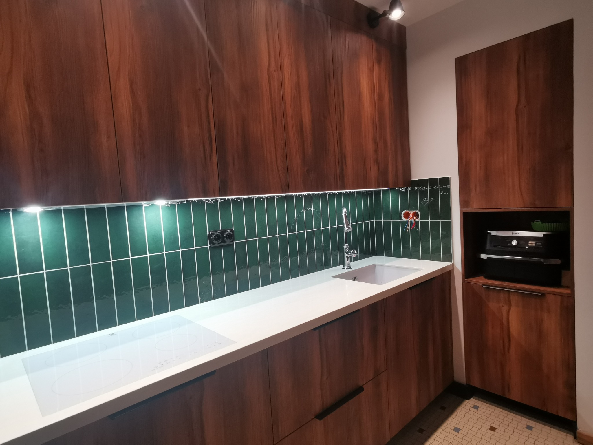 Nowoczesna kuchnia z drewnianymi frontami, białym blatem i zielonymi płytkami ściennymi w stylu retro, uzupełniona o białą płytę indukcyjną i armaturę ze stali nierdzewnej.