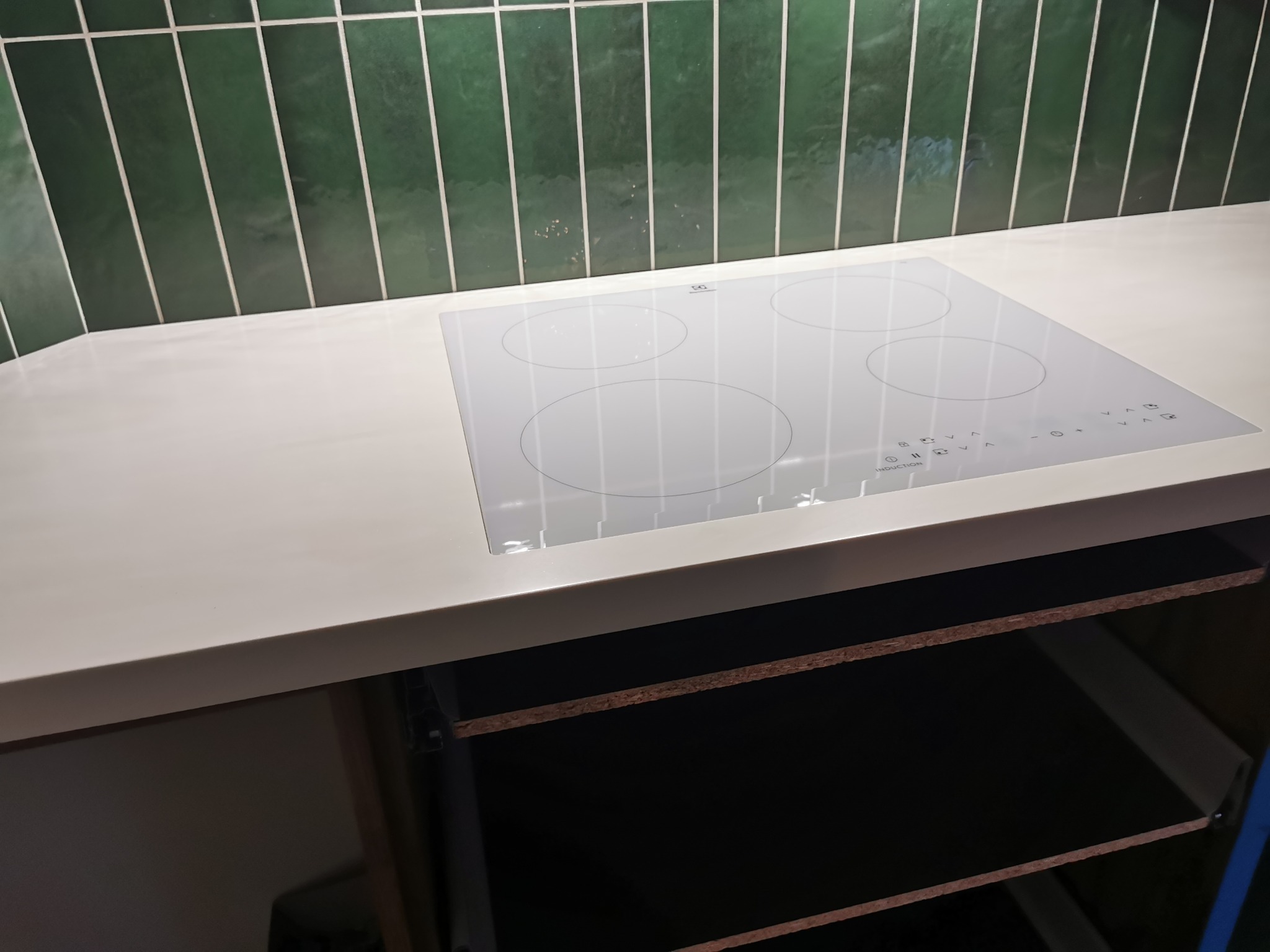 Biała płyta indukcyjna Electrolux wkomponowana w jasny blat kuchenny, z zielonymi, pionowymi płytkami w tle, zamontowana w kuchni w Zielonce.