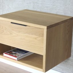 ADM - Wisząca szafka nocna z jasnego drewna dębowego z szufladą z czarnym uchwytem, na półce książka, tło stanowi surowa ściana.