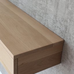 ADM - Minimalistyczna szafka wisząca z litego drewna dębowego, z pojedynczą szufladą, zamontowana na tle ściany o surowym, betonowym wykończeniu.