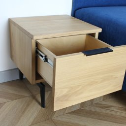 ADM - Wisząca szafka nocna z jasnego drewna z wysuniętą szufladą na metalowych prowadnicach i czarnym uchwytem, na tle niebieskiej sofy i podłogi w jodełkę.