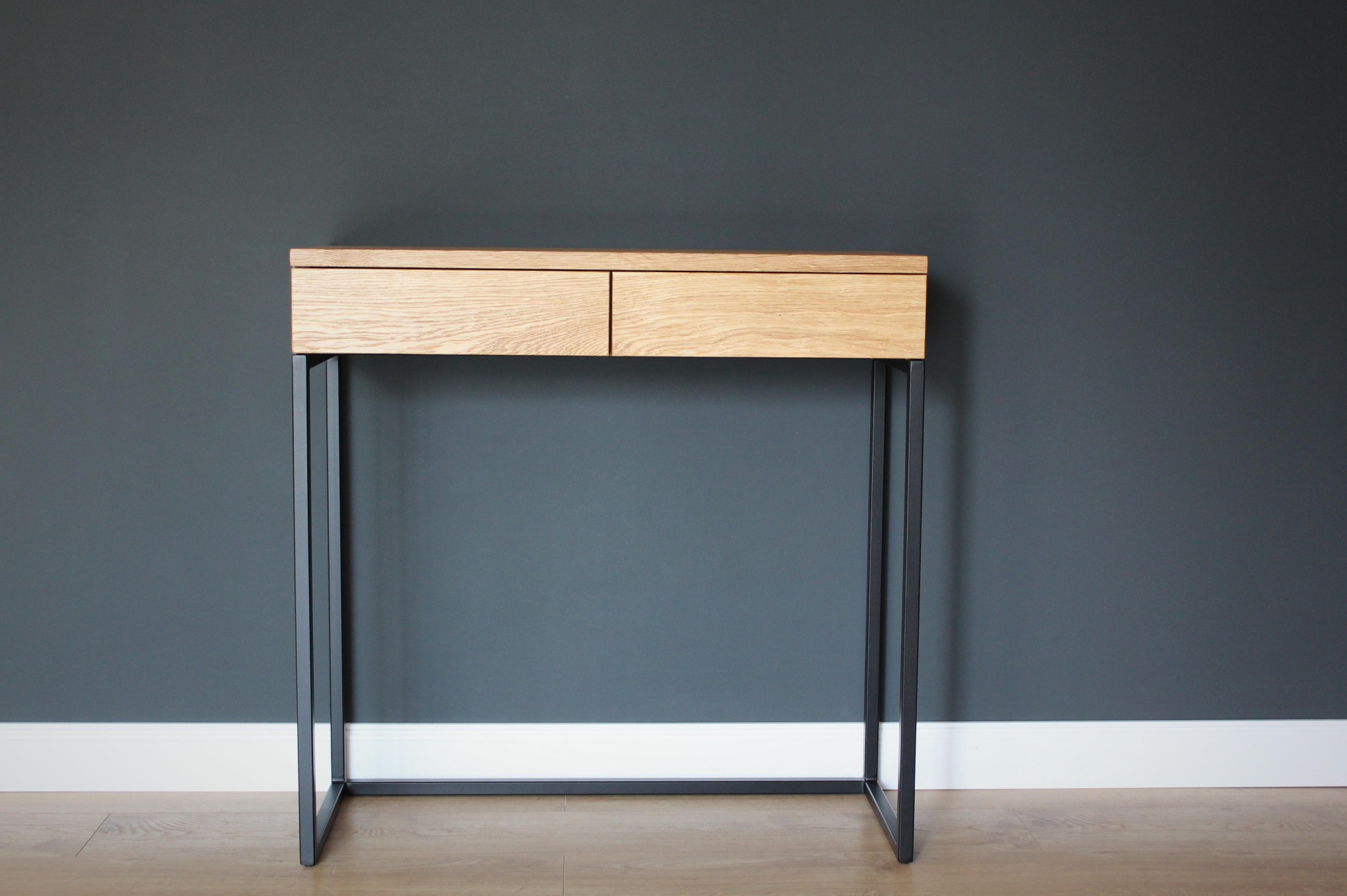 Minimalistyczny stolik konsola z dwoma szufladami z jasnego drewna i czarnymi metalowymi nogami na tle ciemnoszarej ściany i jasnej podłogi.