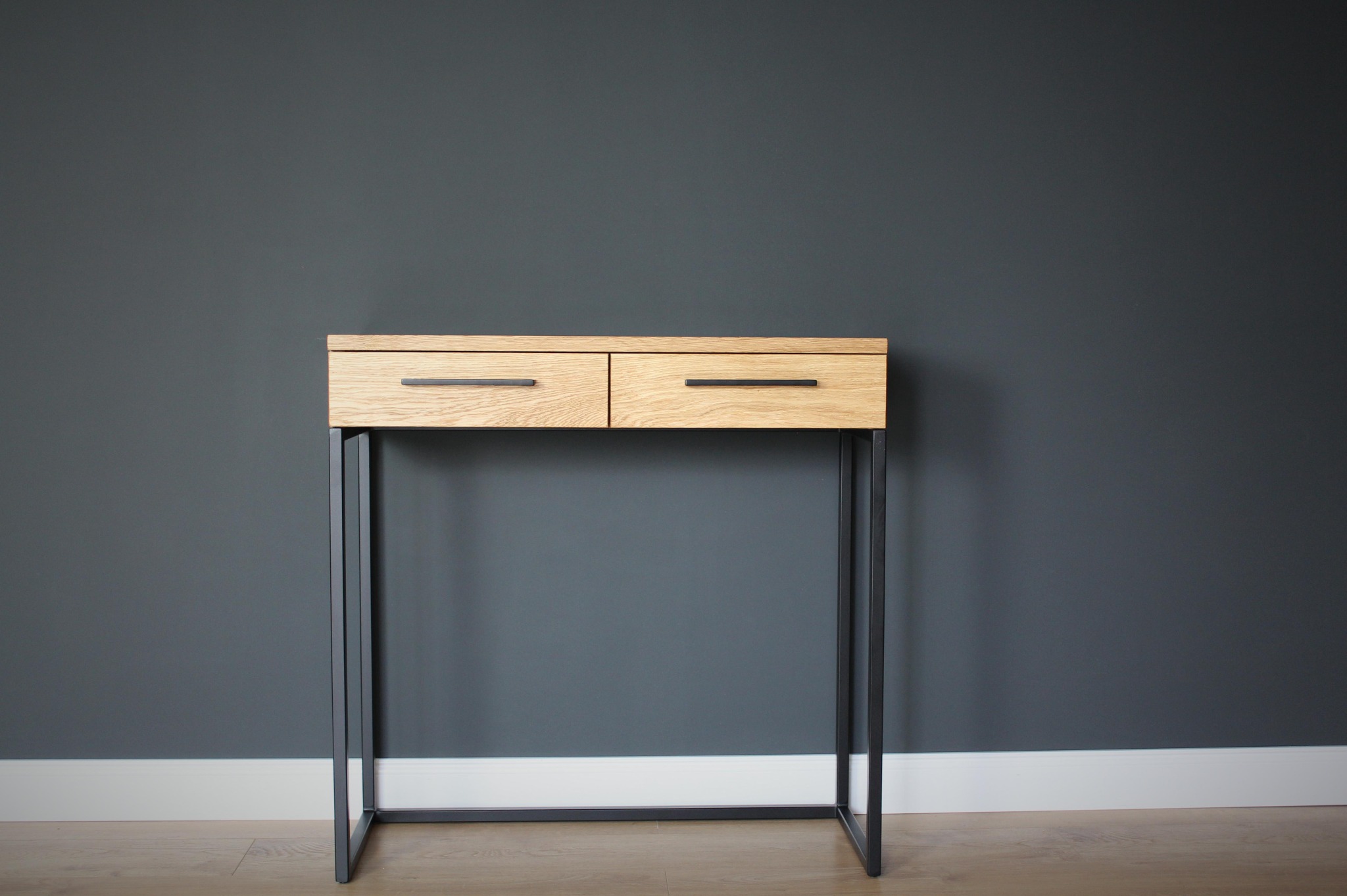 Konsola z dwoma szufladami z jasnego drewna na czarnych, metalowych nogach, na tle szarej ściany z białą listwą przypodłogową.