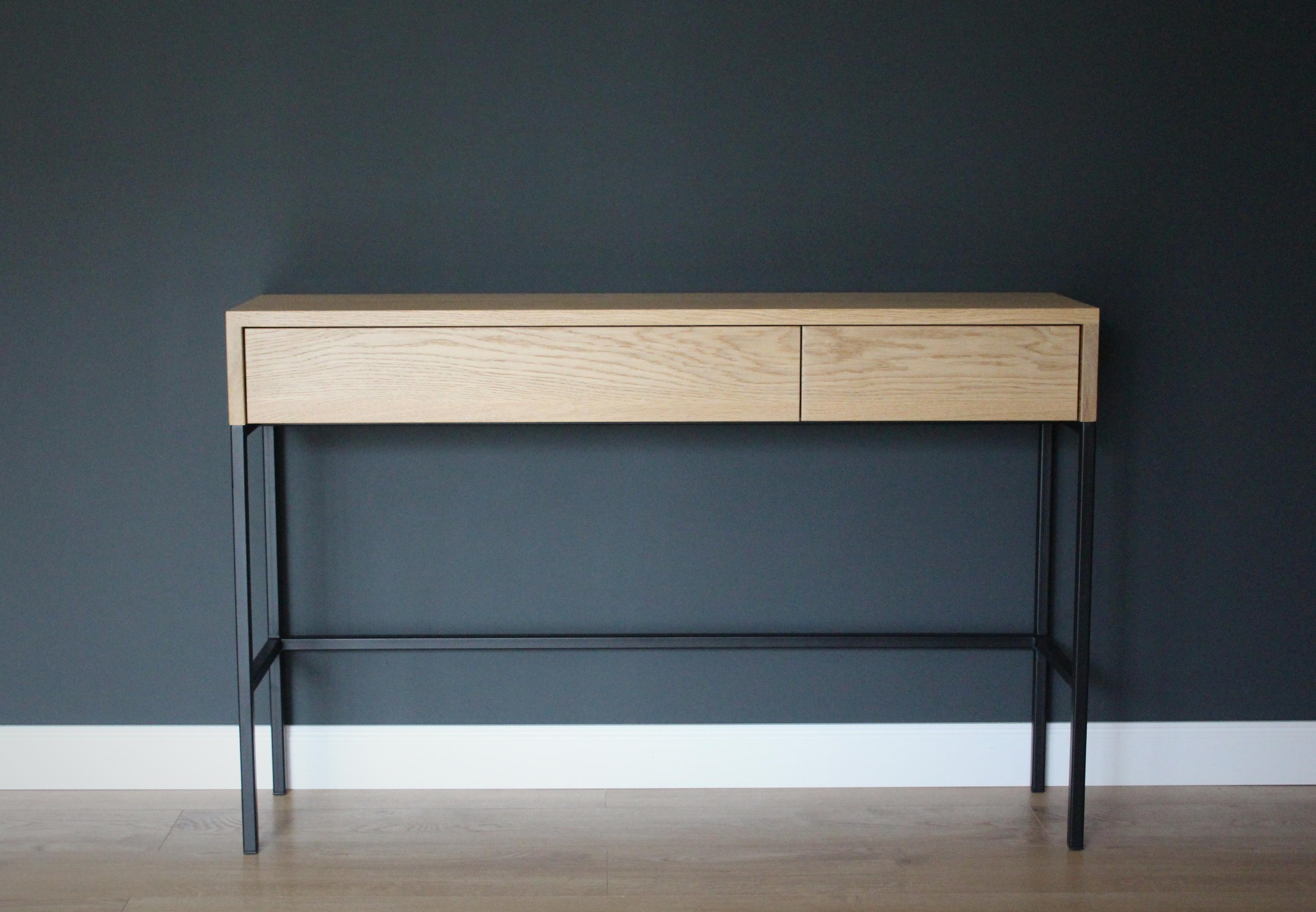 Biurko z jasnego drewna z dwiema szufladami, osadzone na czarnych, metalowych nogach, na tle ciemnoszarej ściany i jasnej podłogi.