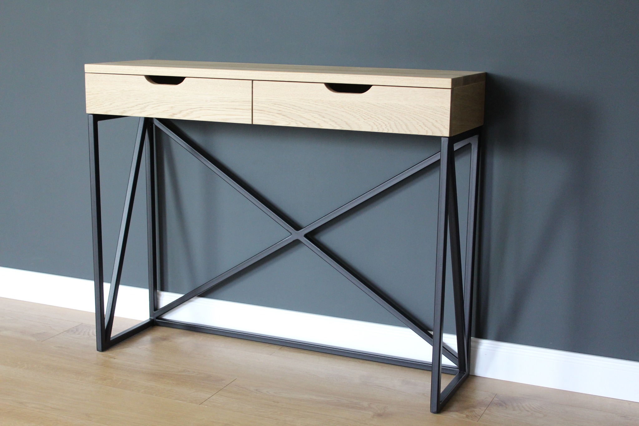 Konsoleta z dwoma szufladami z jasnego drewna na czarnych, metalowych nogach w kształcie litery X, ustawiona na drewnianej podłodze przy szarej ścianie.