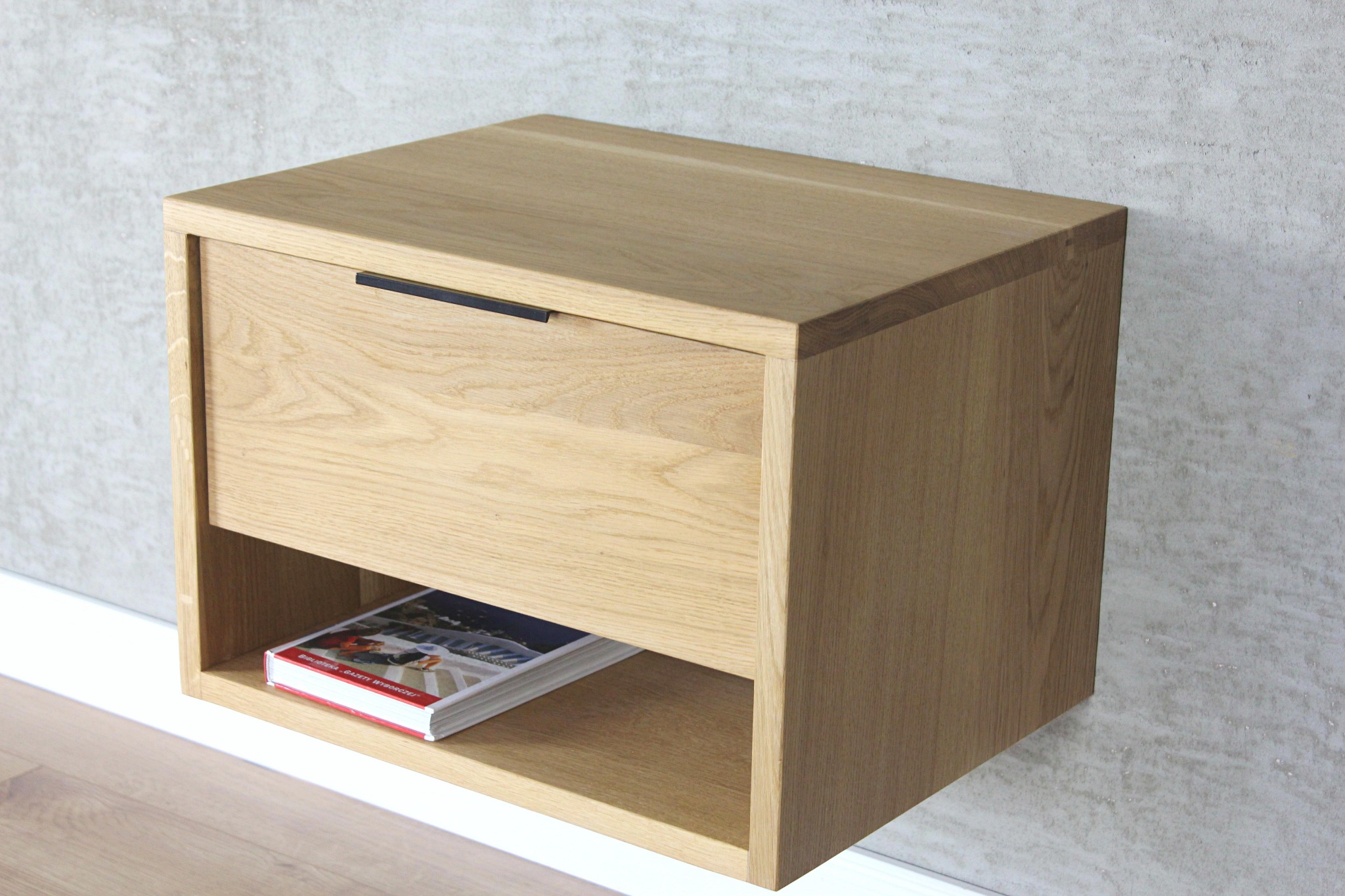 Wisząca szafka nocna z jasnego drewna dębowego z szufladą z czarnym uchwytem, na półce książka, tło stanowi surowa ściana.