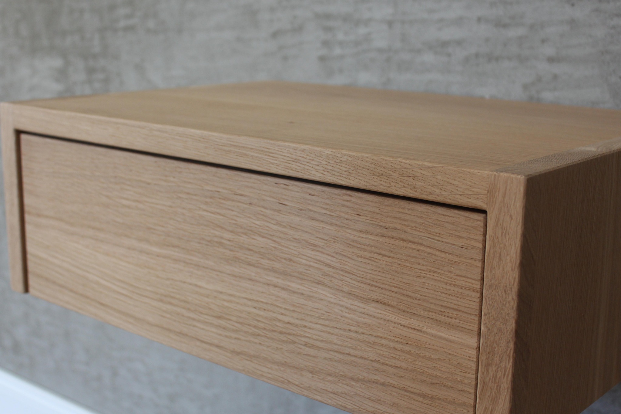 Półka nocna z litego drewna dębowego, zawieszona na ścianie o szarej, betonowej fakturze, widoczne usłojenie drewna i minimalistyczny design.