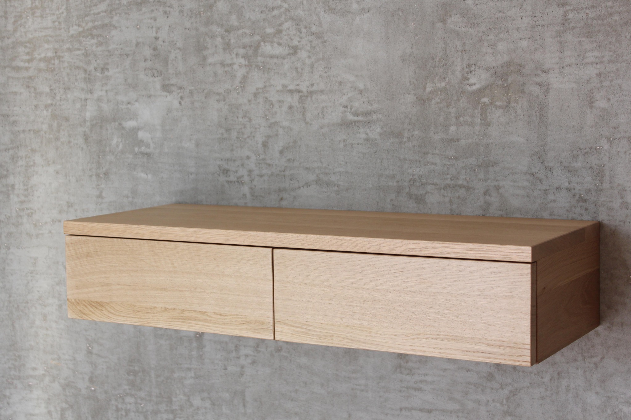 Drewniana szafka wisząca z dwiema szufladami, zamontowana na surowej, betonowej ścianie.