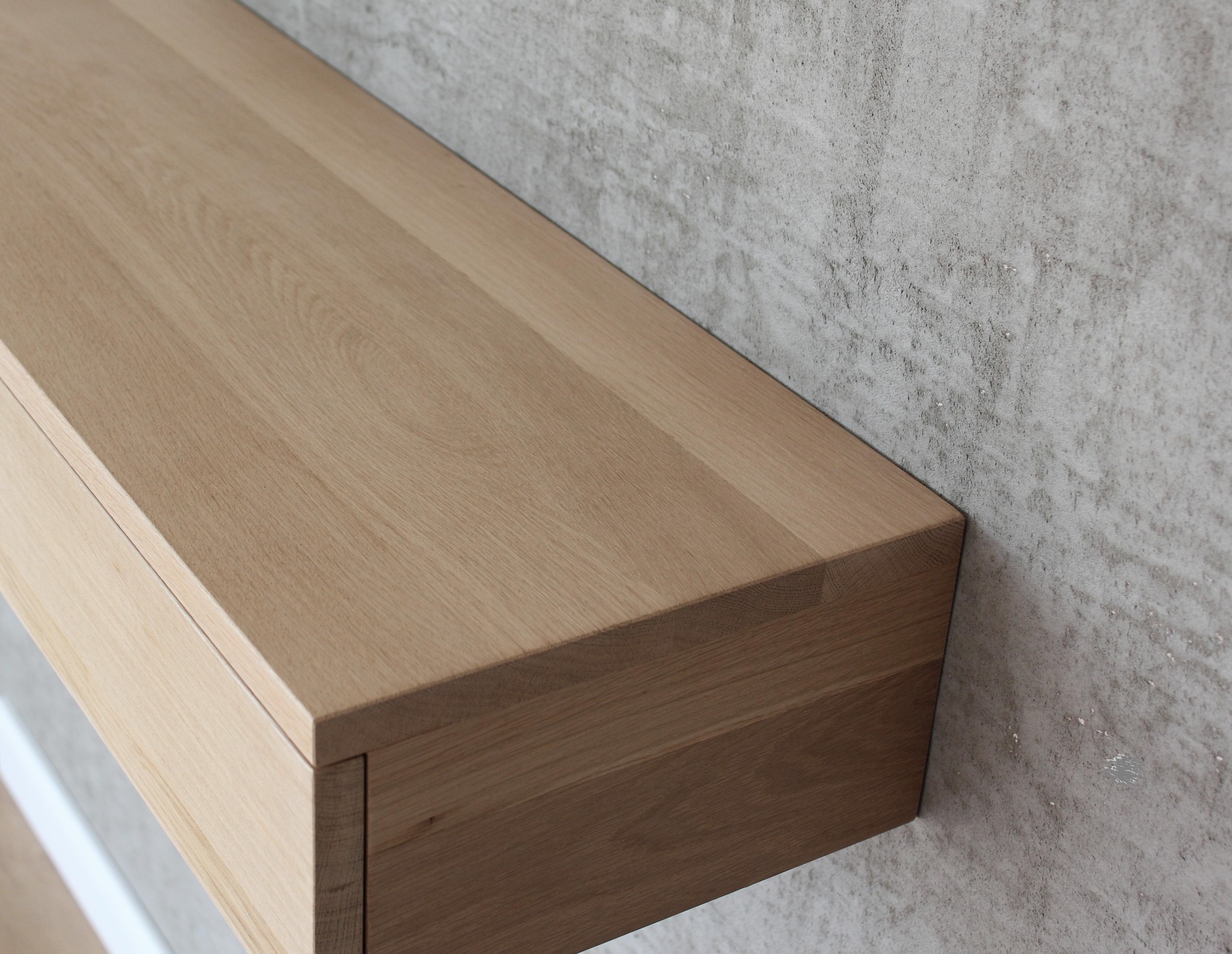 Minimalistyczna szafka wisząca z litego drewna dębowego, z pojedynczą szufladą, zamontowana na tle ściany o surowym, betonowym wykończeniu.