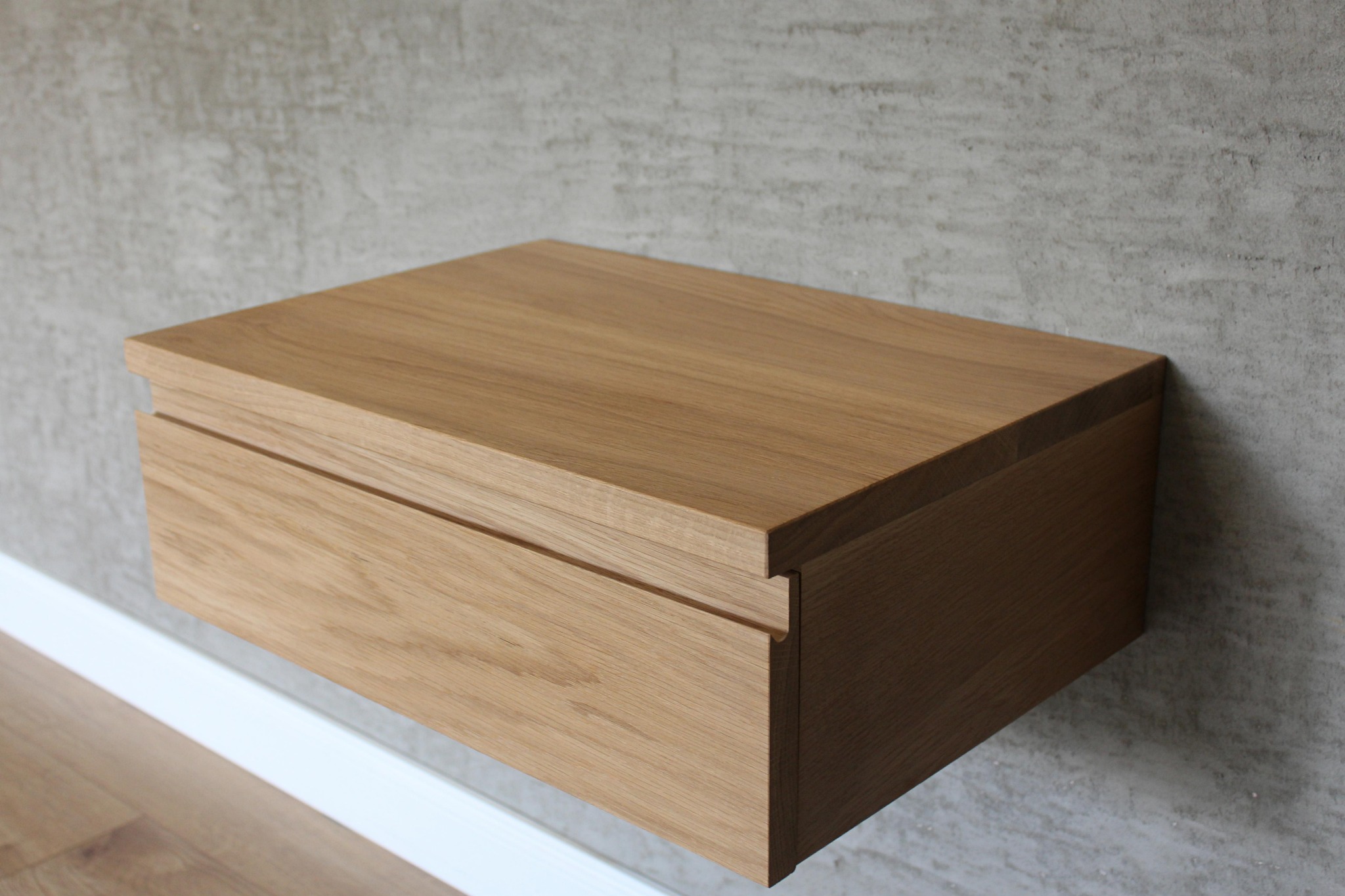 Drewniana szafka nocna z szufladą, zawieszona na ścianie o fakturze betonu, minimalistyczny design.