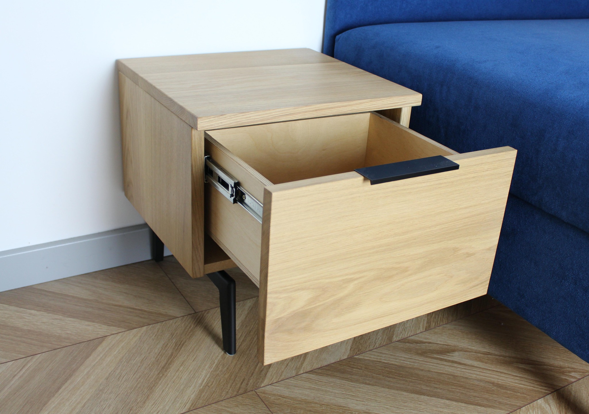 Wisząca szafka nocna z jasnego drewna z wysuniętą szufladą na metalowych prowadnicach i czarnym uchwytem, na tle niebieskiej sofy i podłogi w jodełkę.