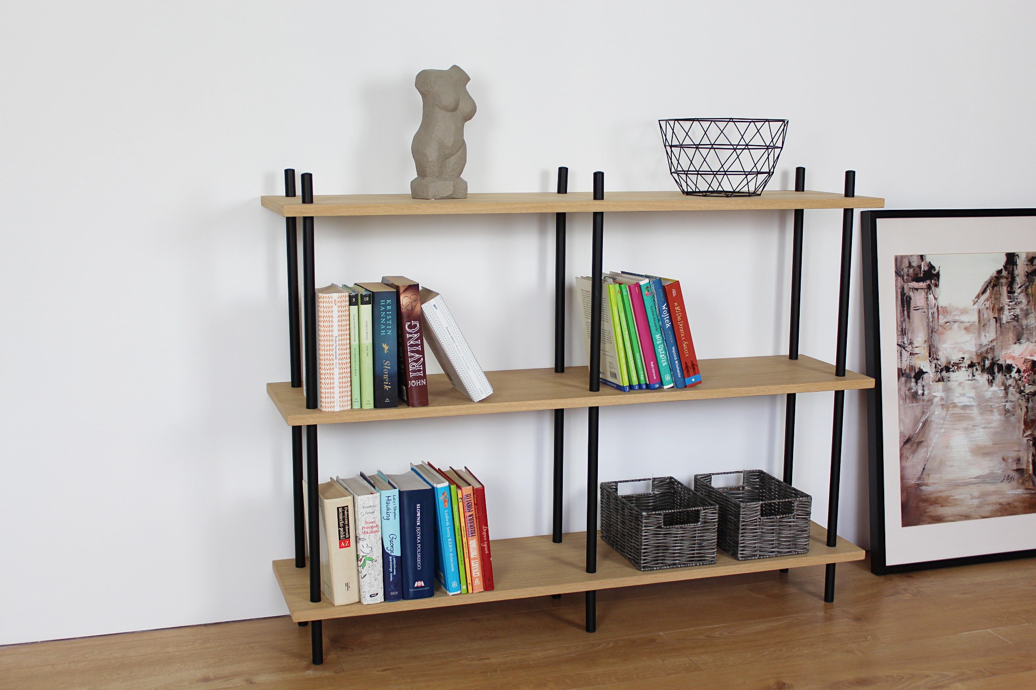 Regał z trzema półkami z jasnego drewna i czarnymi metalowymi nogami, na półkach książki, rzeźba popiersia, koszyk druciany i dwa wiklinowe pojemniki, obok obraz w czarnej ramie.
