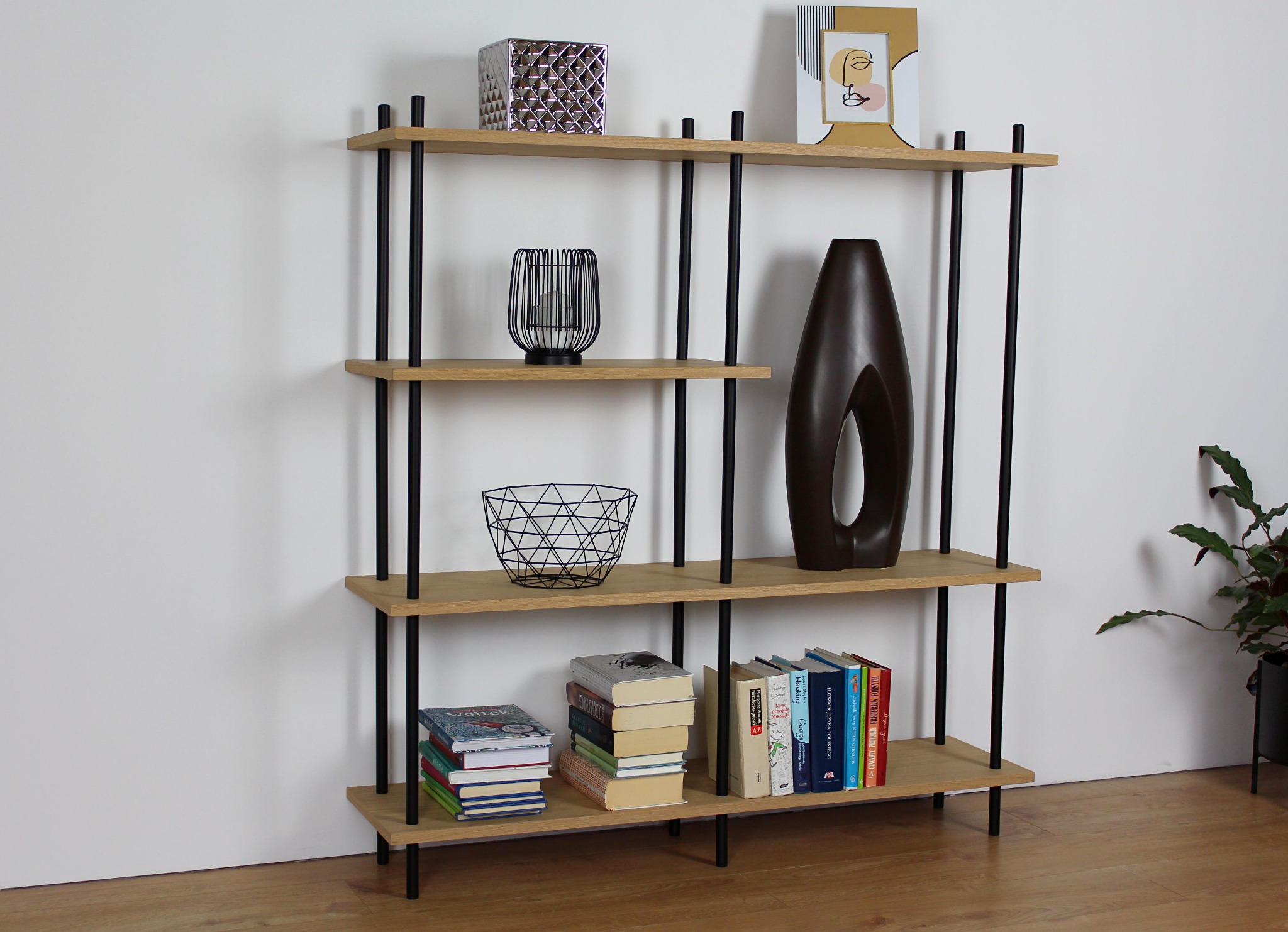 Regał z jasnego drewna i czarnych metalowych nóżek, na którym znajdują się książki, wazon i ozdobne przedmioty, ustawiony na drewnianej podłodze przy białej ścianie.