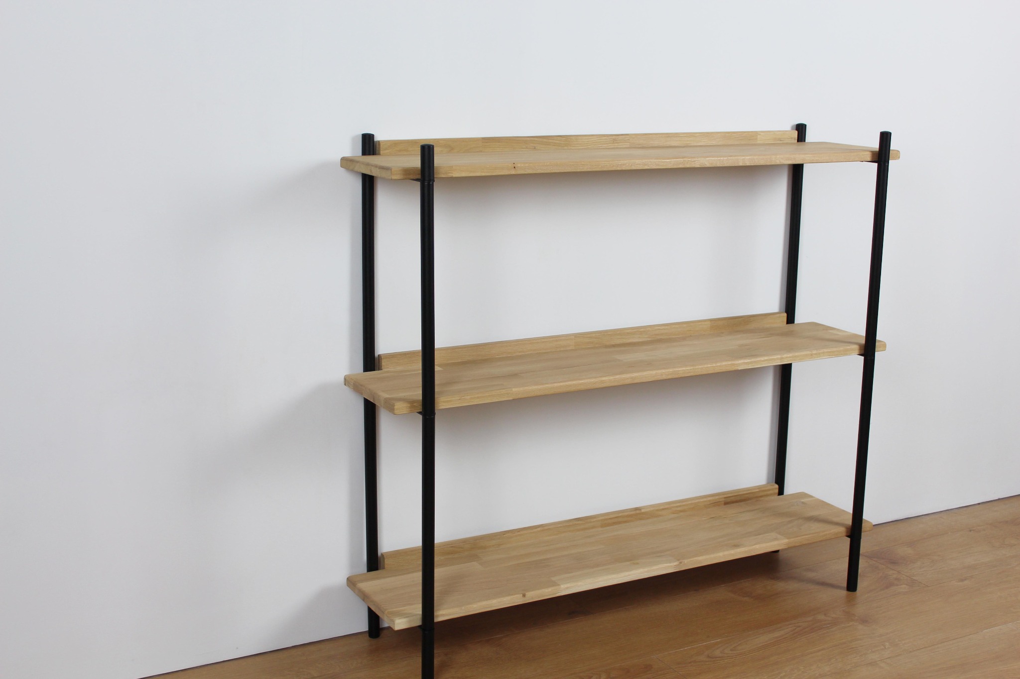 Trzypoziomowy regał z jasnego drewna z czarnymi metalowymi nogami, ustawiony na drewnianej podłodze przy białej ścianie.